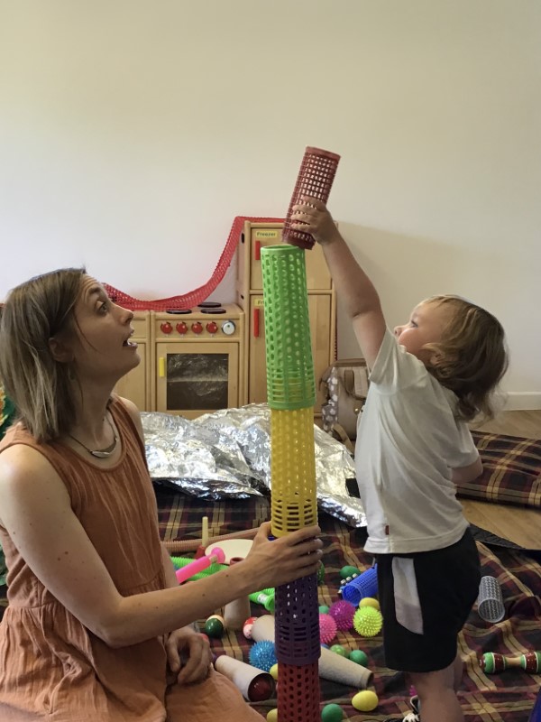 Charlotte Jolly and a child are sitting on rugs on the floor. They are building a tower from coloured,stackable, plastic cylinders.