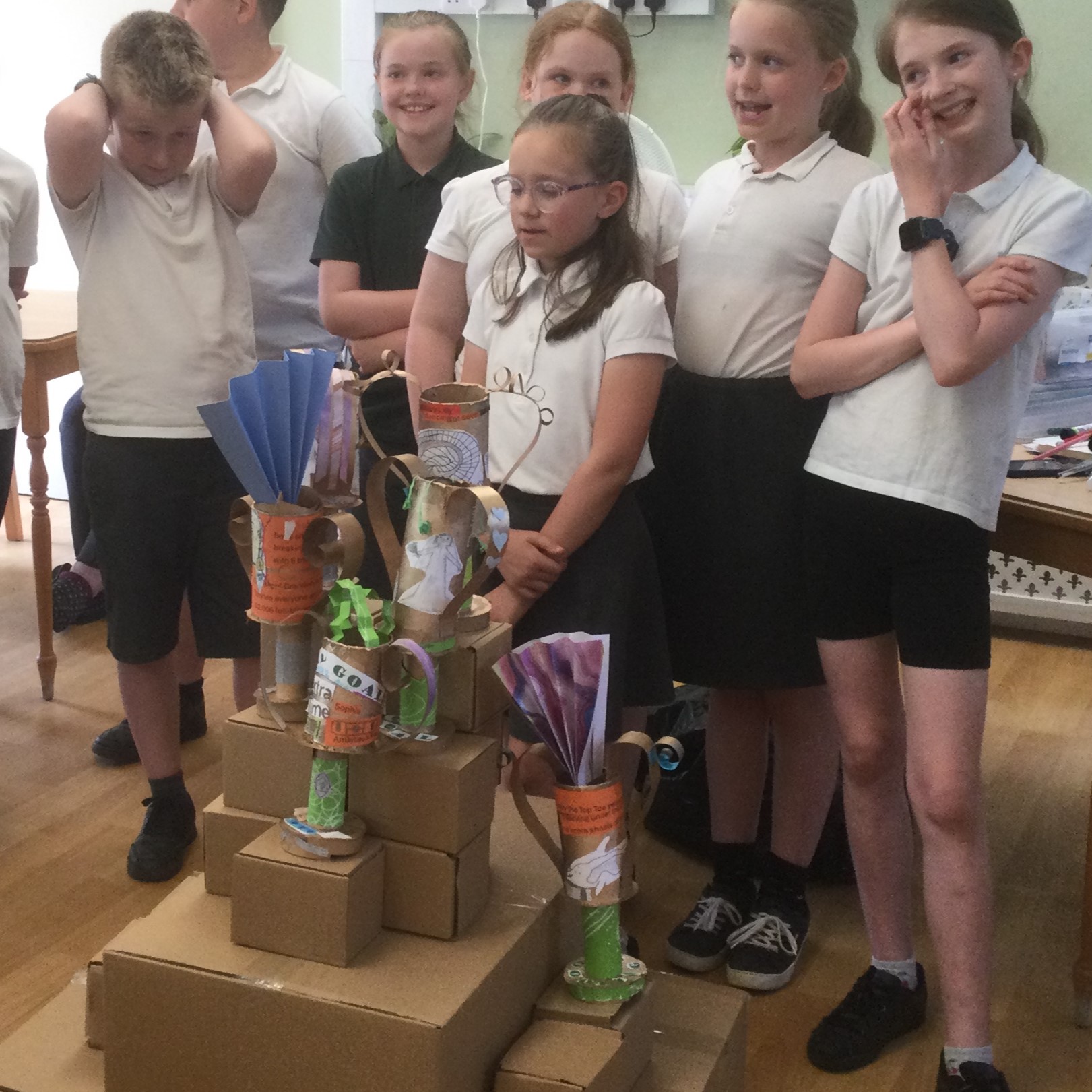 A group of school children stand behind a tower (podium) of cardboard boxes on which stand a variety of cardboard trophies