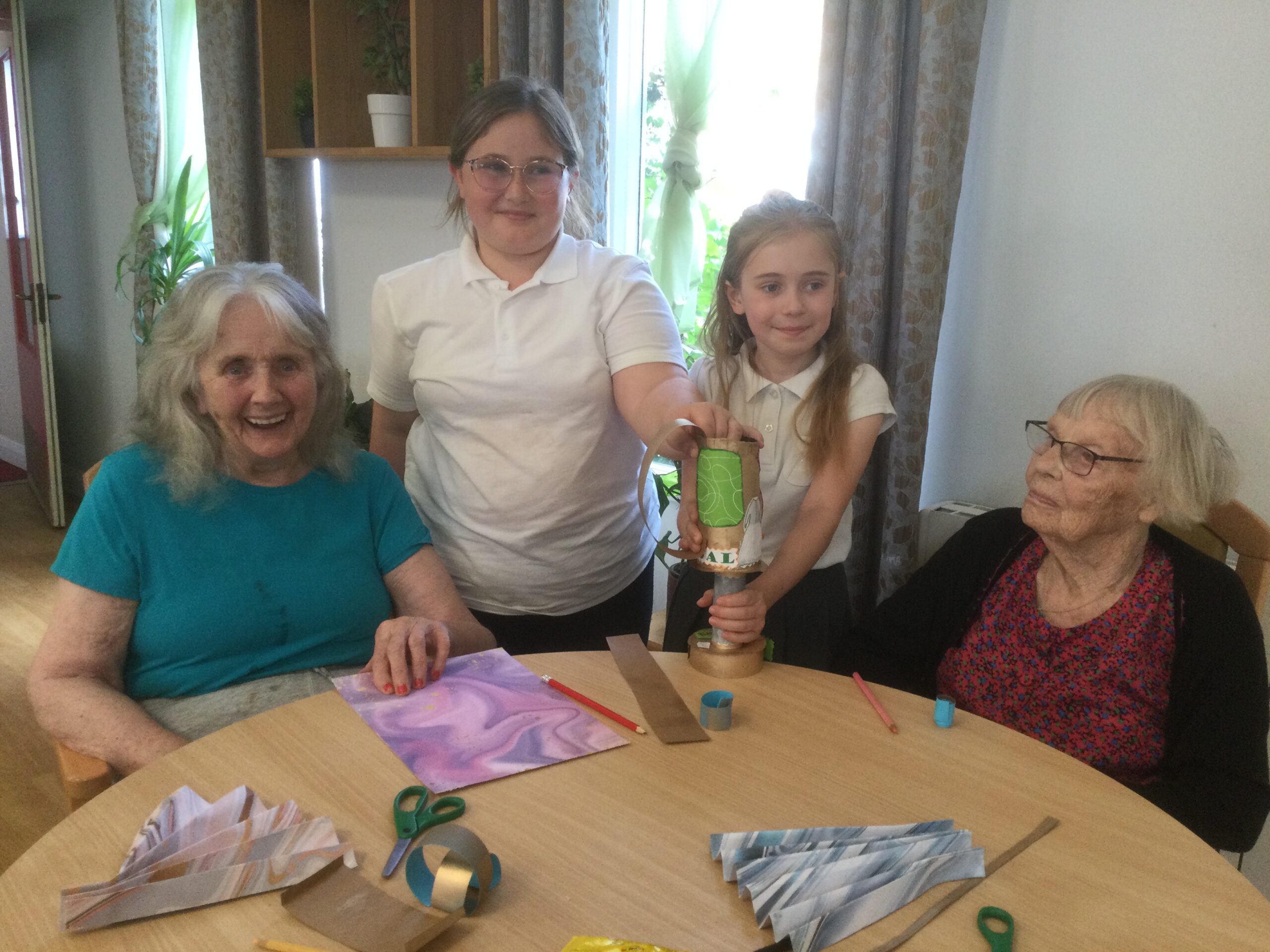 Two school girls stand beside two elderly residents, holding a handmade trophy