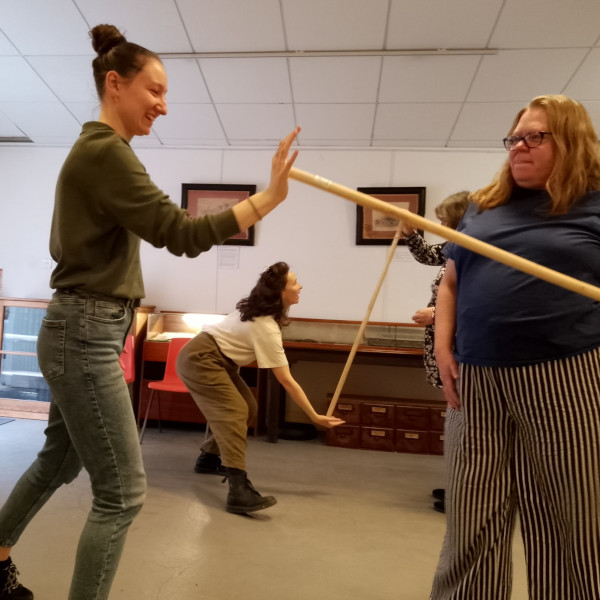 This photograph shows people in a workshop working in pairs to explore movement. Each pair has a pole which is held between them by each person pressing the palm of one hand against either end of the pole. You can see that the pairs are trying different movements while keeping the pole from falling by maintaining an even pressure on each end of the pole. The participants are concentrating and laughing a lot too.