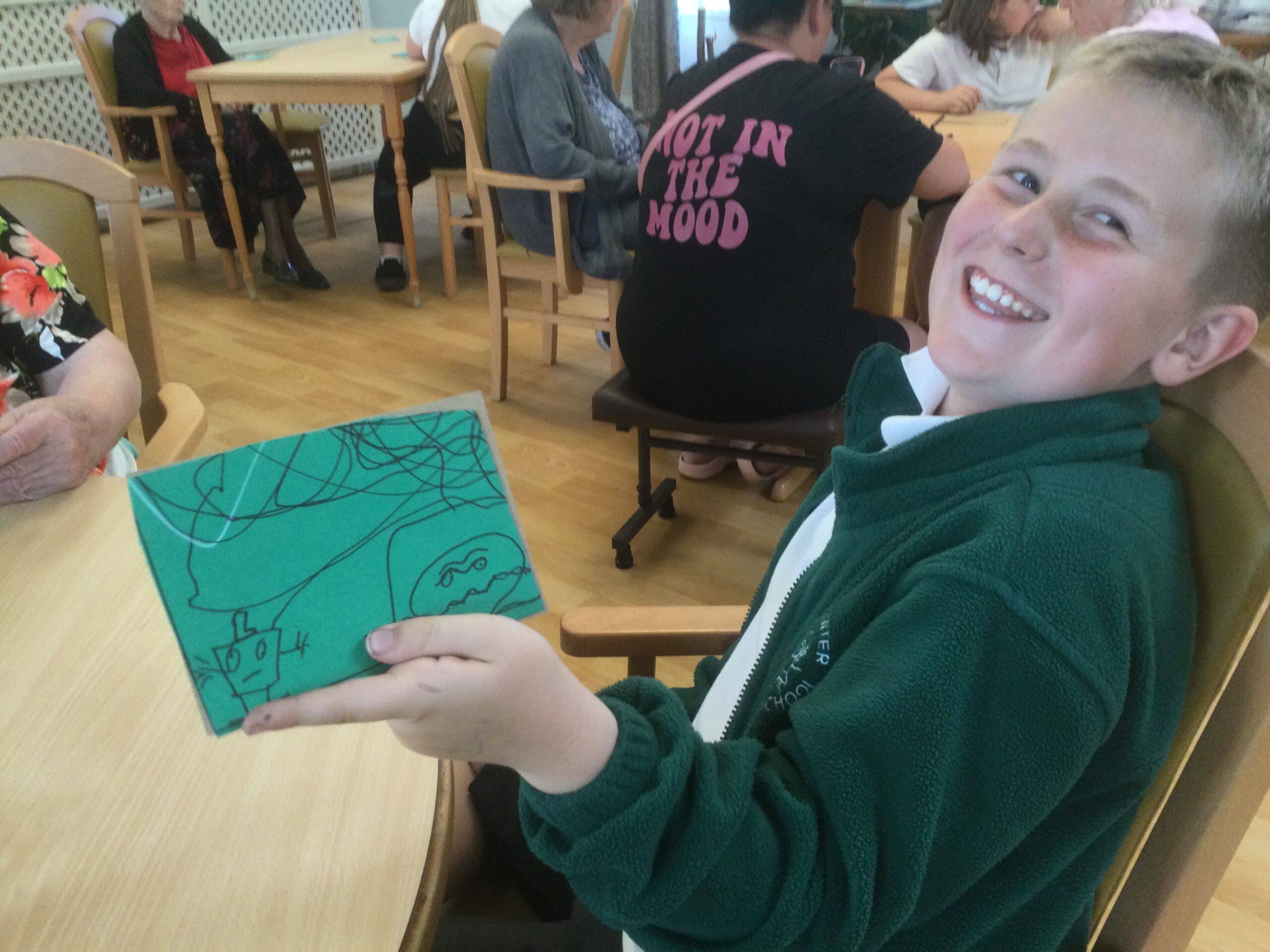 A school boy smiles broadly at the camera whilst showing a black and white line drawing on a green background. Other figures are seated in the background