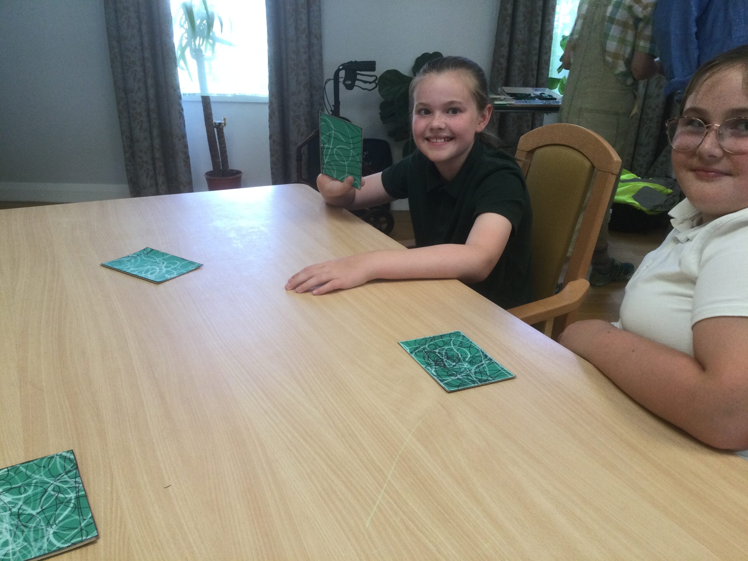 Two school girls, one holding up a green card with black and white line drawings on it. On the table in front of them are three further green cards, each with white and black line drawing