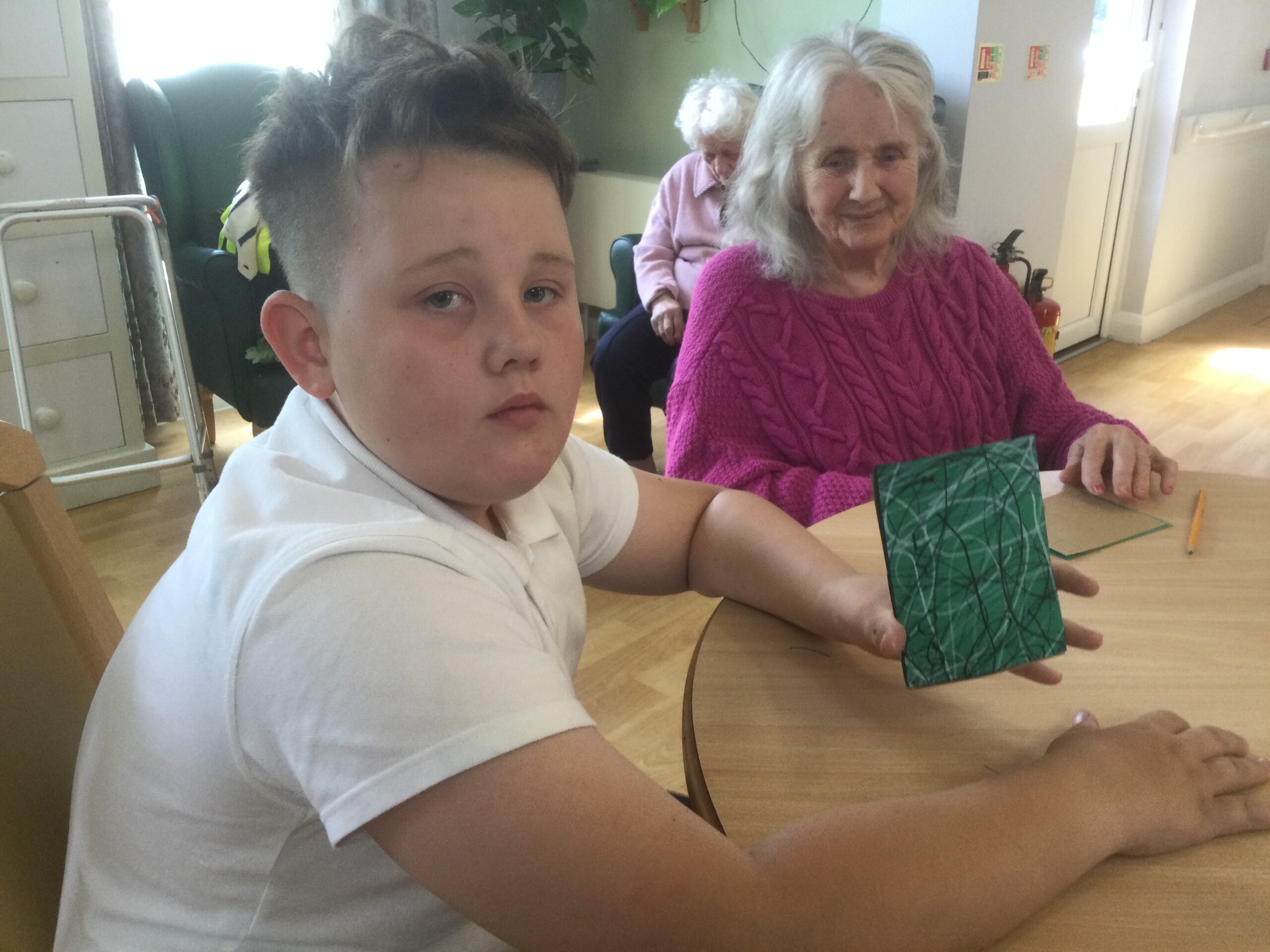 A school boy holds up a green postcard with black and white line drawings whilst a woman dressed in a pink jumper sits smiling down at the table