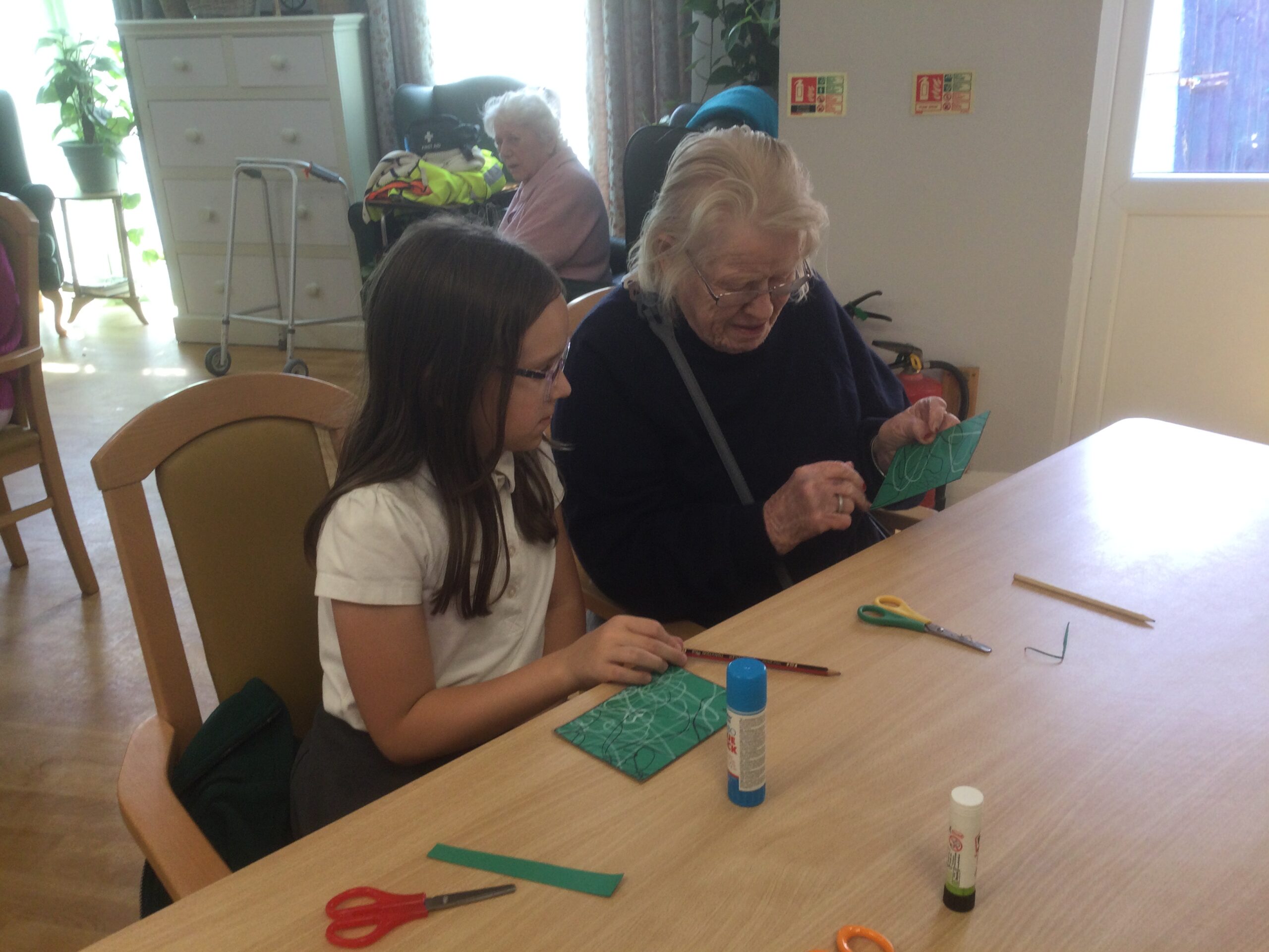 A school girl seated beside and adult, looking at the green cards with line drawings on them. On the table in front of them are scissors and glue sticks