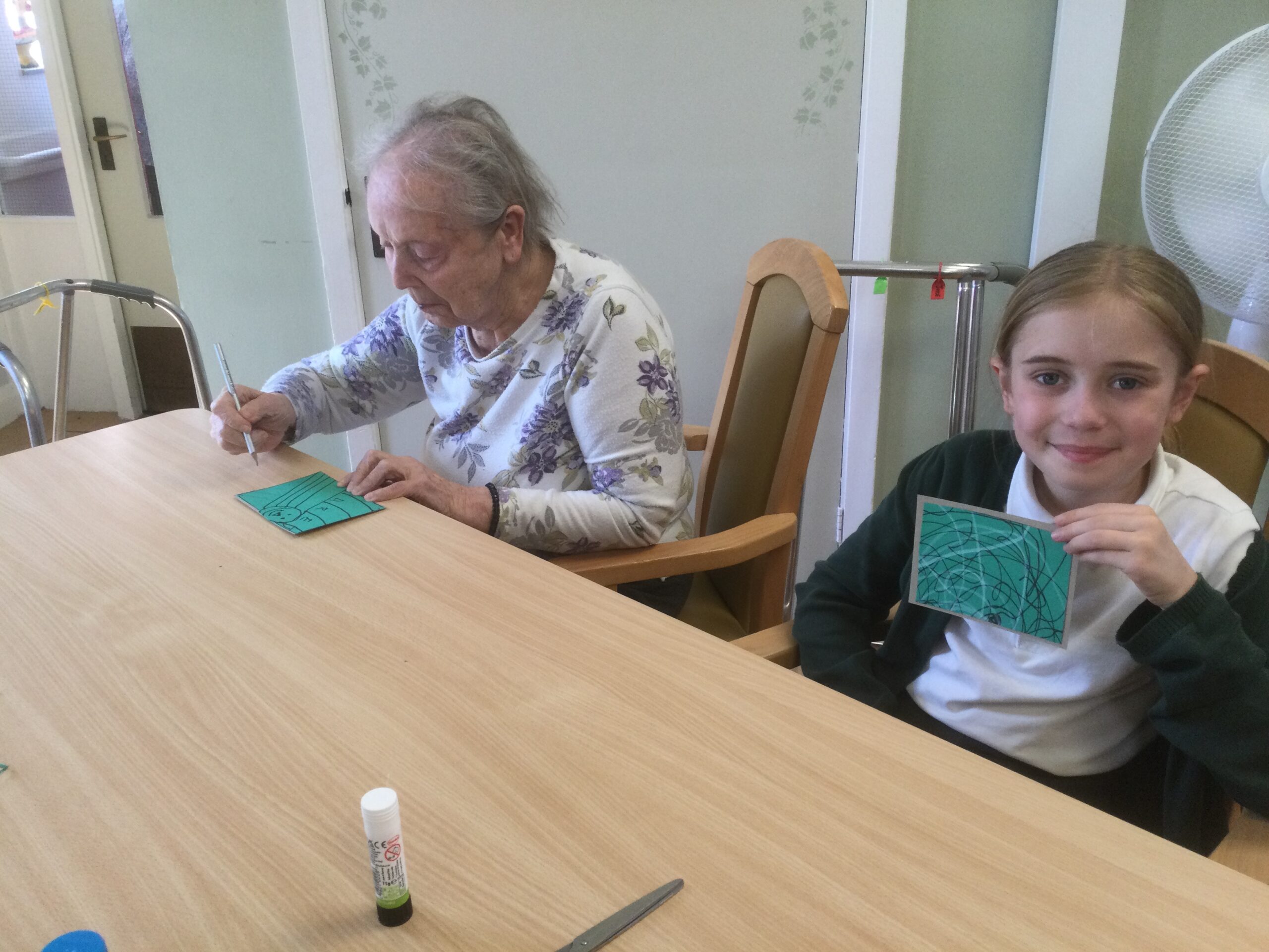 A school girl holds up a green card, with white and black line drawings on it. Beside her is seated a woman who is adding lines to her card