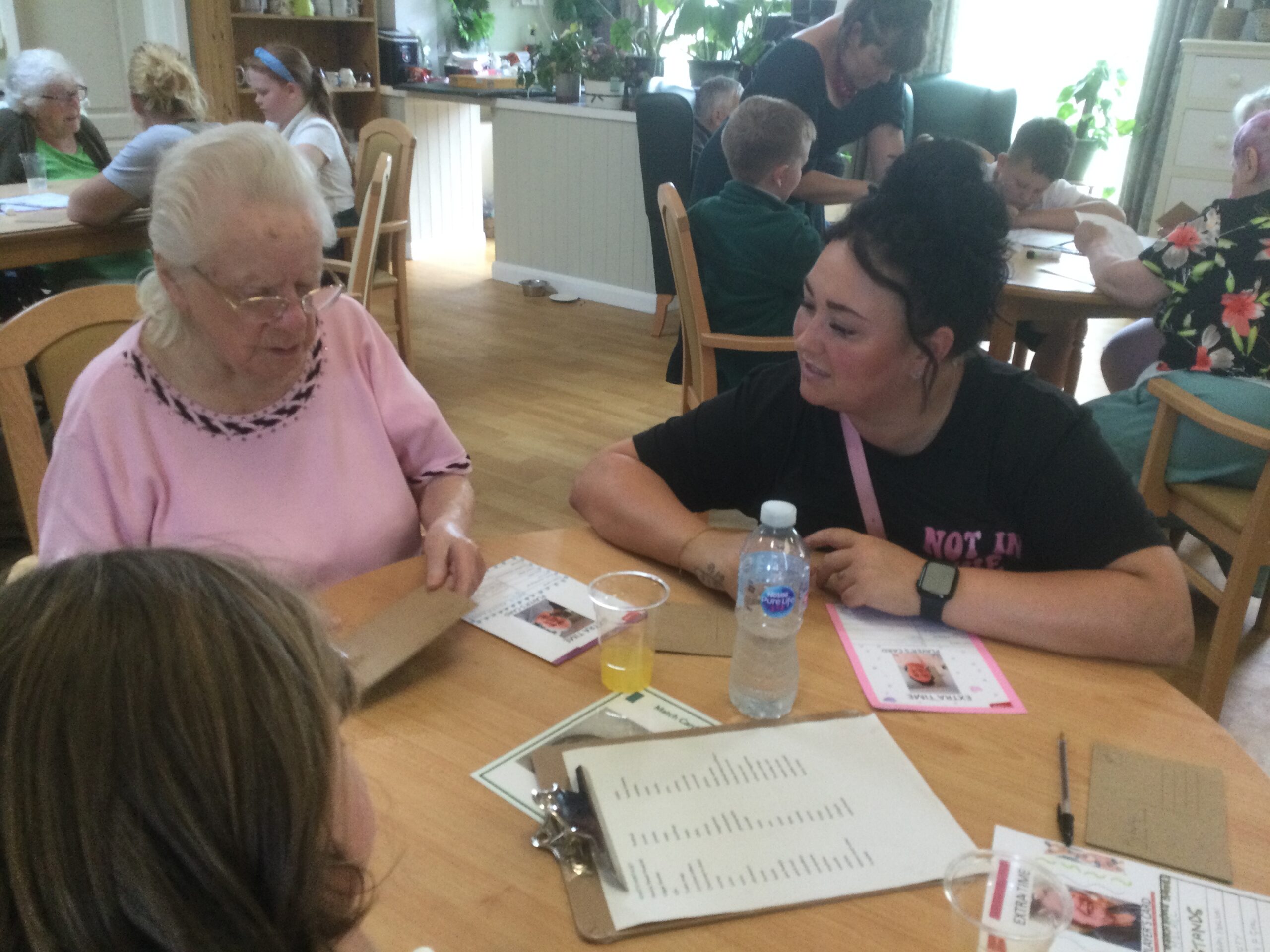 An elderly lady dressed in pink is holding a brown postcard whilst another woman with a black T-shirt sits beside her, chatting. Other people are in the background