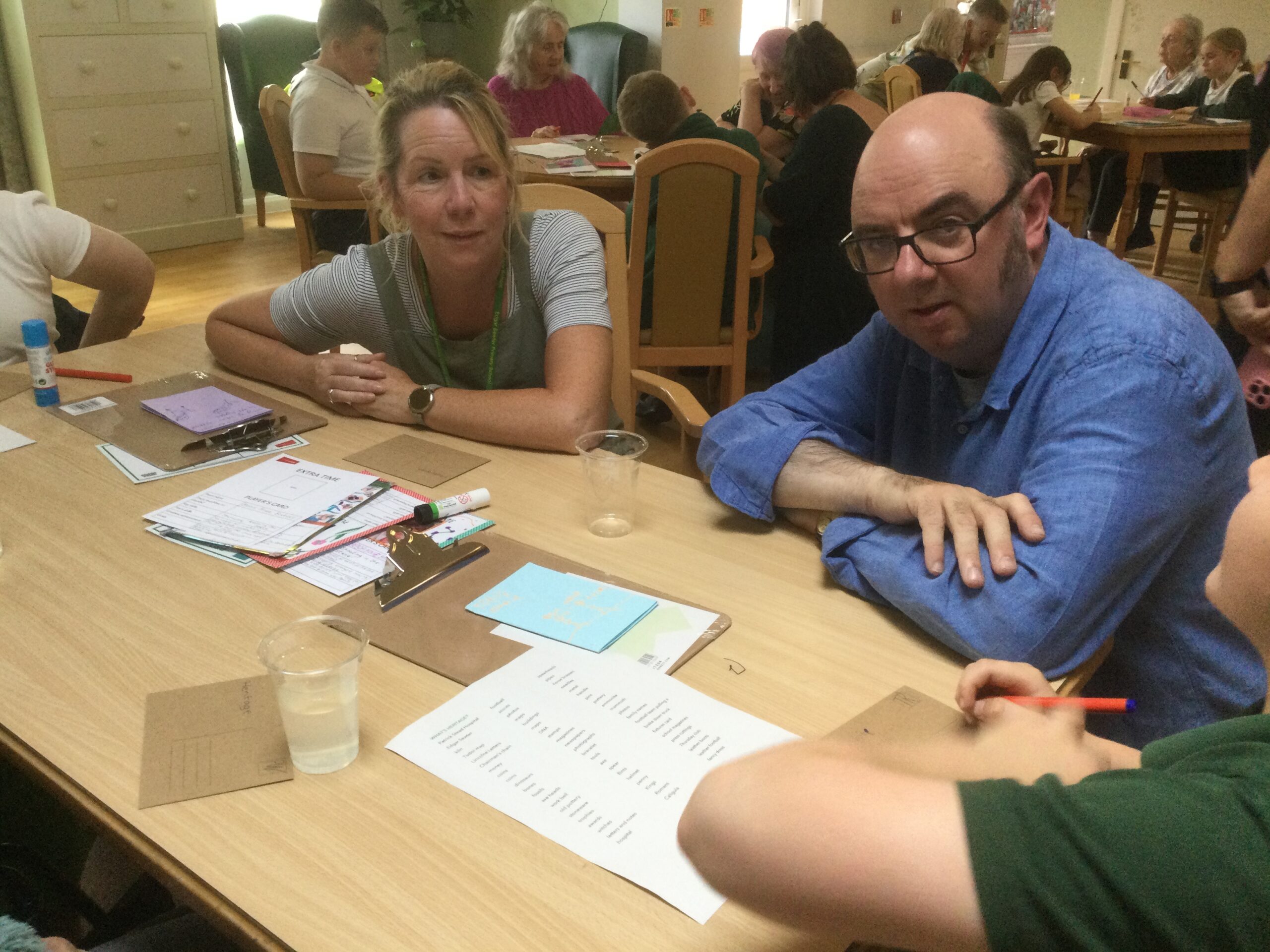 A gentleman in blue shirt leans on a table, with a woman seated to his right and school boy to his left; the table is covered in papers, pens and glue sticks. Several other people are in the background