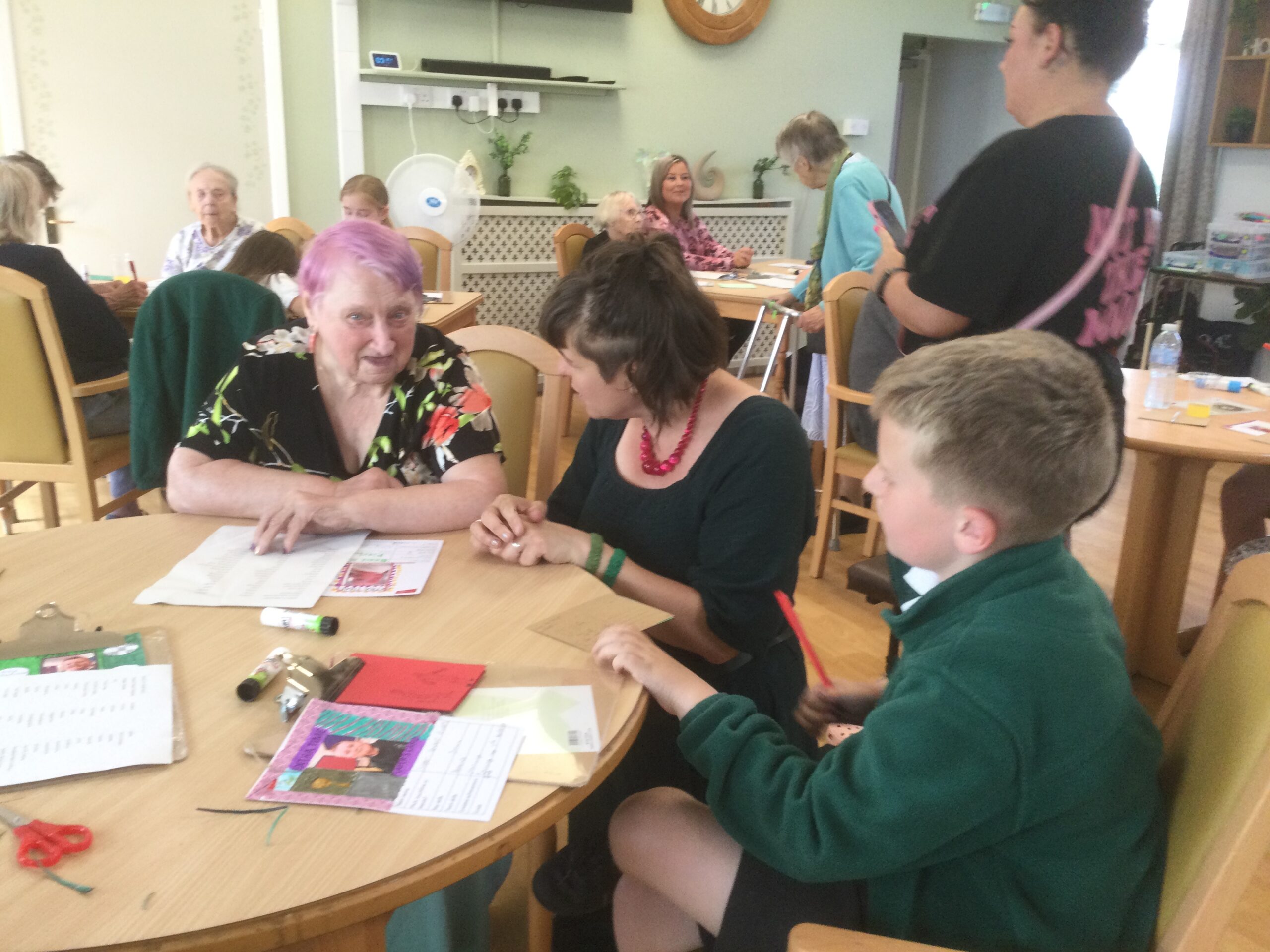 A woman with a big red bead necklace and dark dress kneels beside a woman in a floral frock as the pair chat together. Beside them is a school boy dressed in a thick green fleece and there are other adults in the background