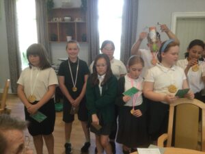 A row of children each wearing a gold medal and holding postcards. One child in the background holds up a trophy