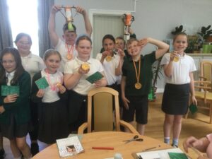 A row of school children holding gold-painted medals, green postcards and, in the background, the trophies