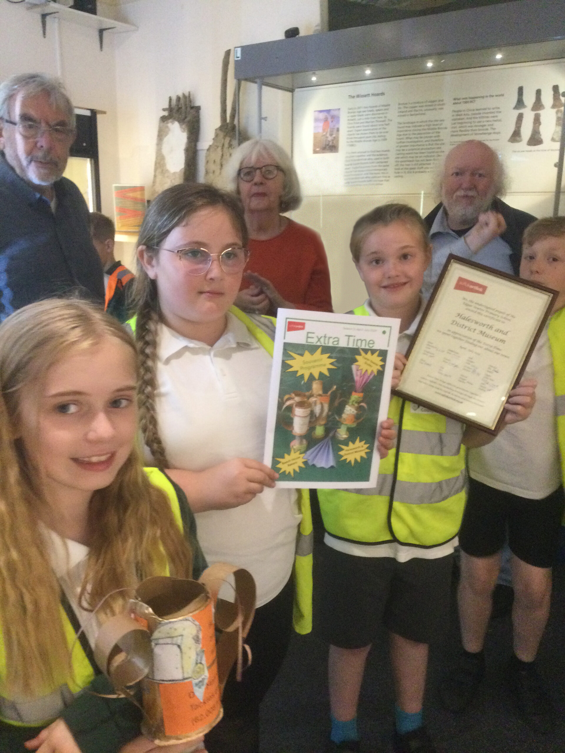 School children holding a framed certificate, a publication and a trophy with a row of three adults standing behind them