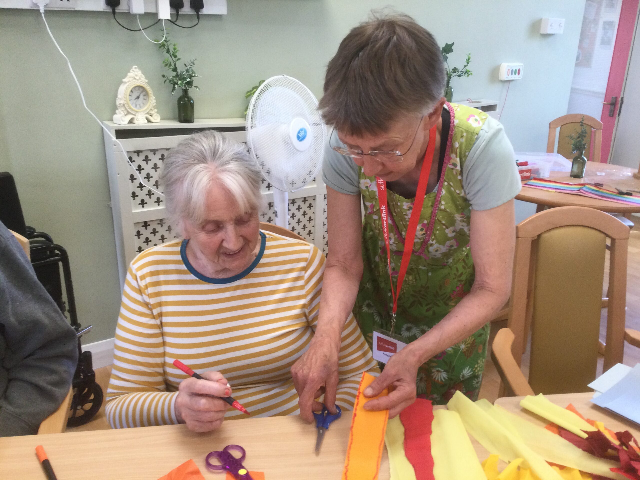 A woman in a white and yellow jersey sits at a table, holding a felt tip pen in her right hand. Beside her stand another woman, picking up a pair of scissors and a piece of yellow tissue paper. There are strips of yellow and red paper on the table in front of them