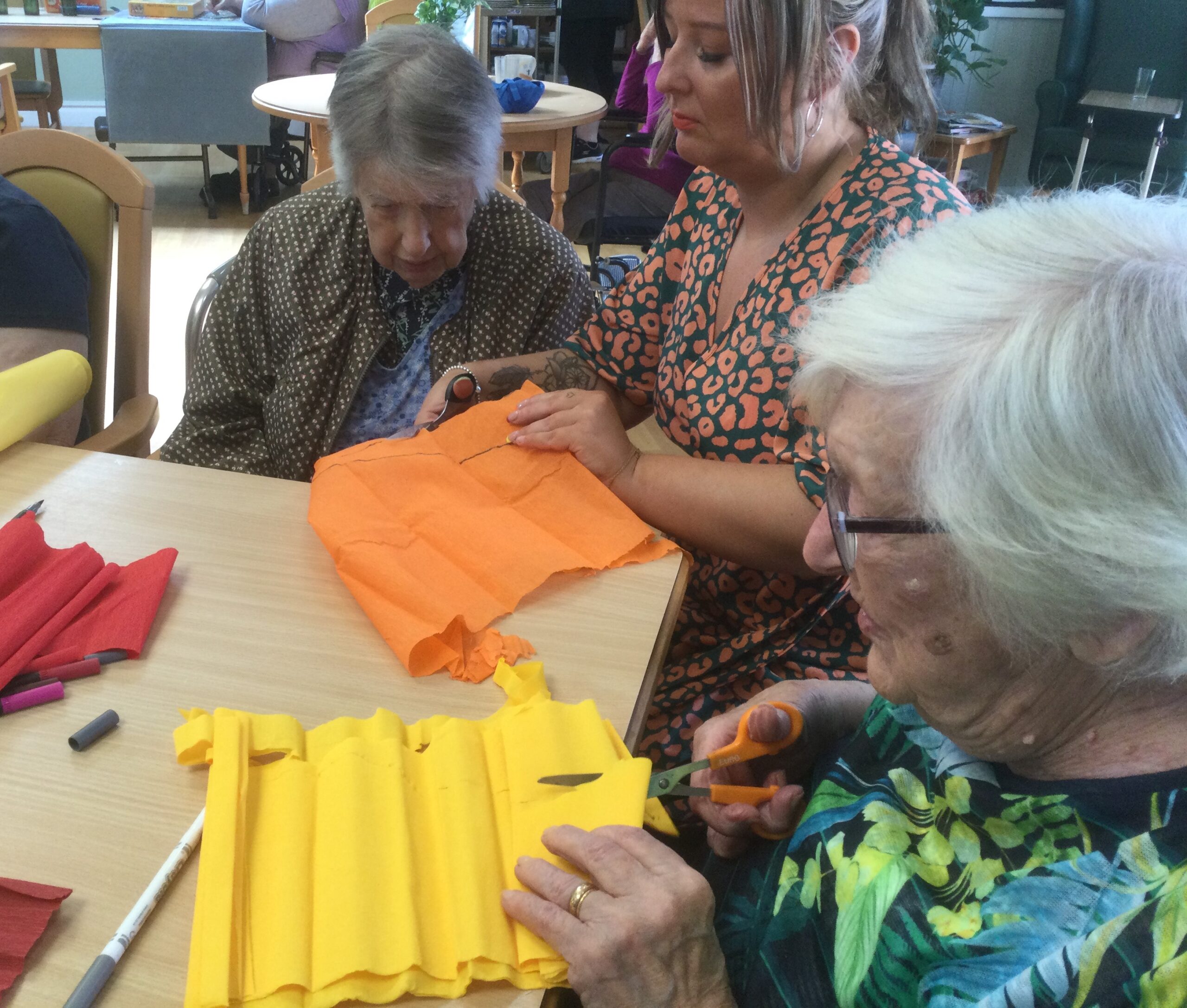 Three women sit at at a table cutting out sheets of orange, yellow and red tissue paper