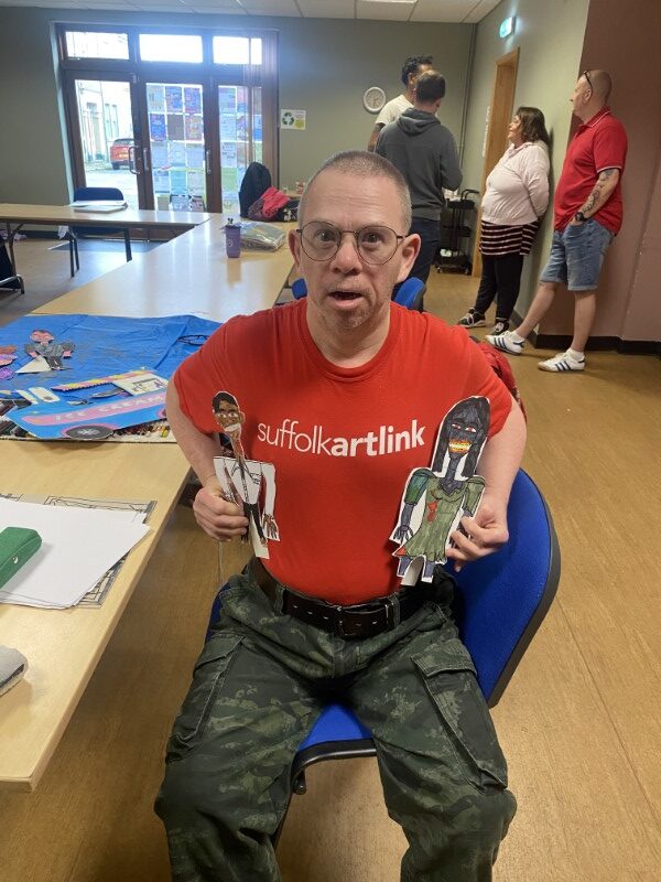 Eric is wearing a red t-shirt with ‘Suffolk Artlink’ emblazoned across the front. He is holding two cutout zombie figures of Mr and Mrs Sunack. Each figure is about 30cms tall.