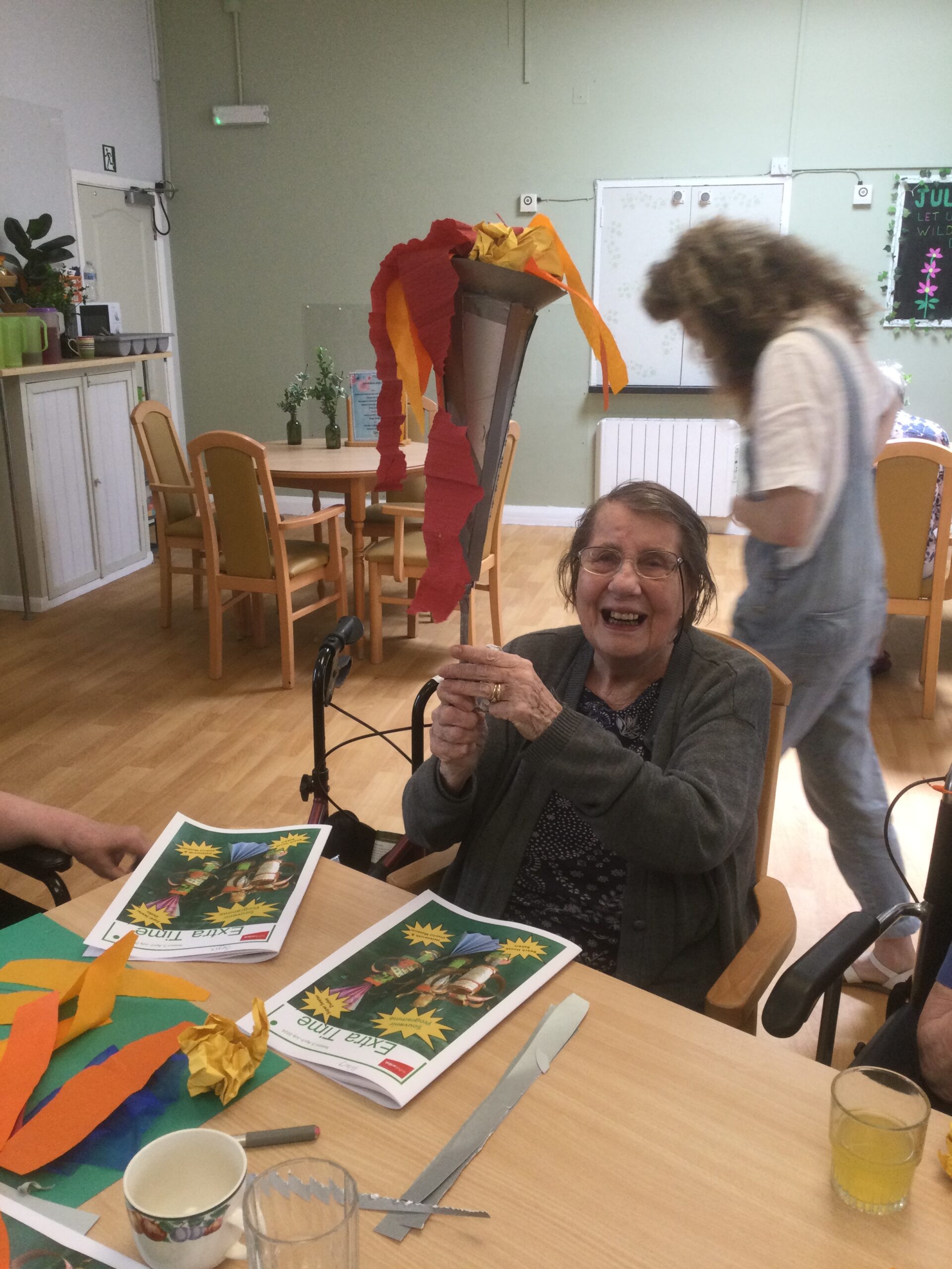 A woman holds up a silver coloured card torch, decorated with red and yellow 'flames. A person in a white T-shirt and denim dungarees walks behind her. On the table are two magazines and some craft materials