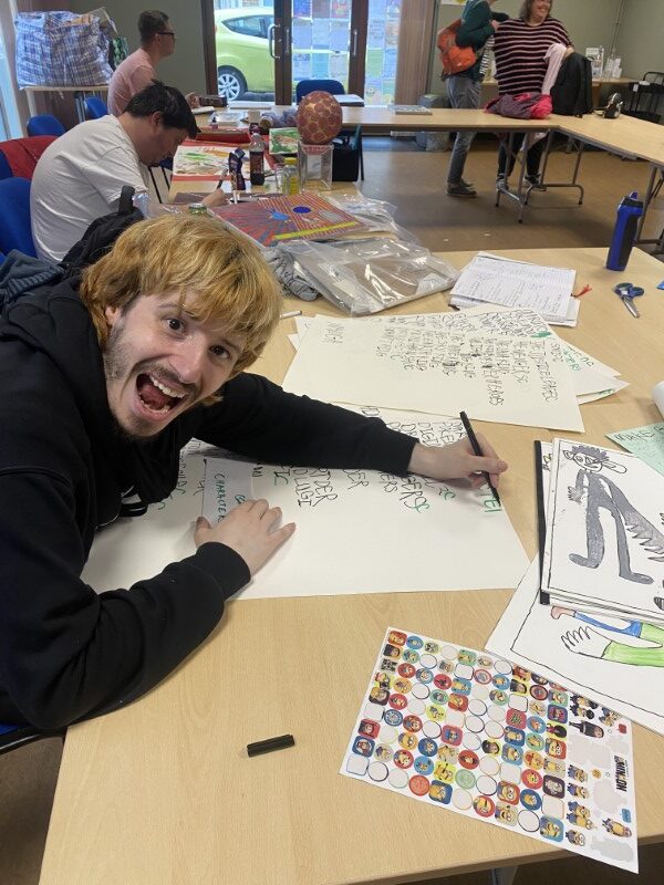 Jake’s face is alight with enthusiasm. He has written words to describe his comic strip scene on a large sheet of paper, but the words are obscured by his arm which is resting across the page.