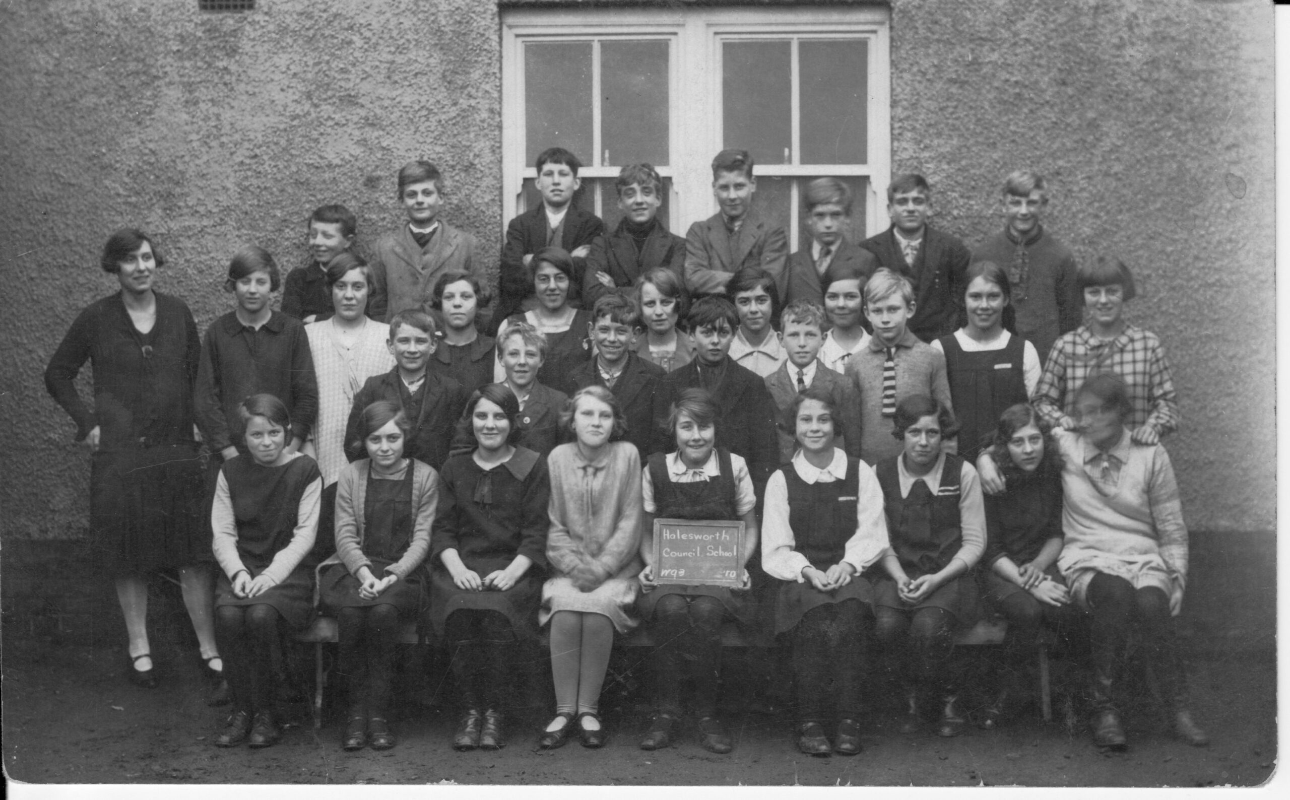 A monochrome photograph from the 1950 showing a row of school children in front of a school wall. A child in the front middle holds a slate on which is written Halesworth Council School