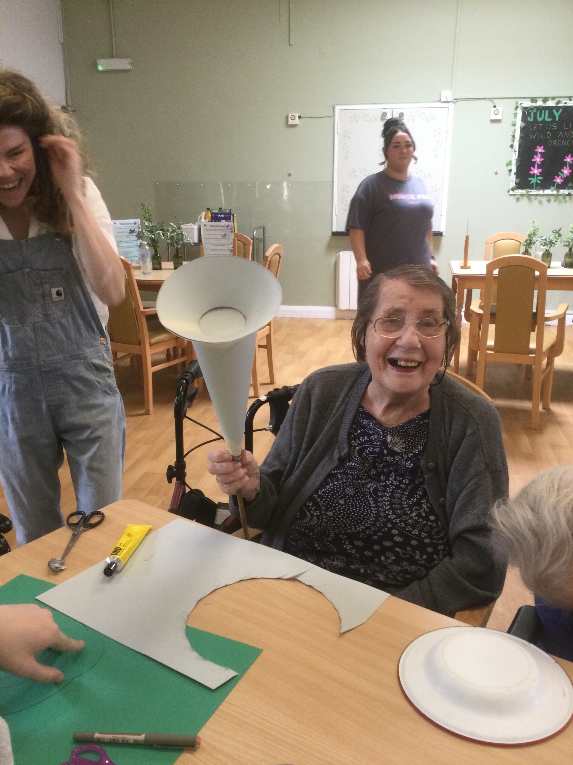 A woman holding a cardboard cone grins happily at the camera. On the table in front of her are pens, a tube of glue and some scissors