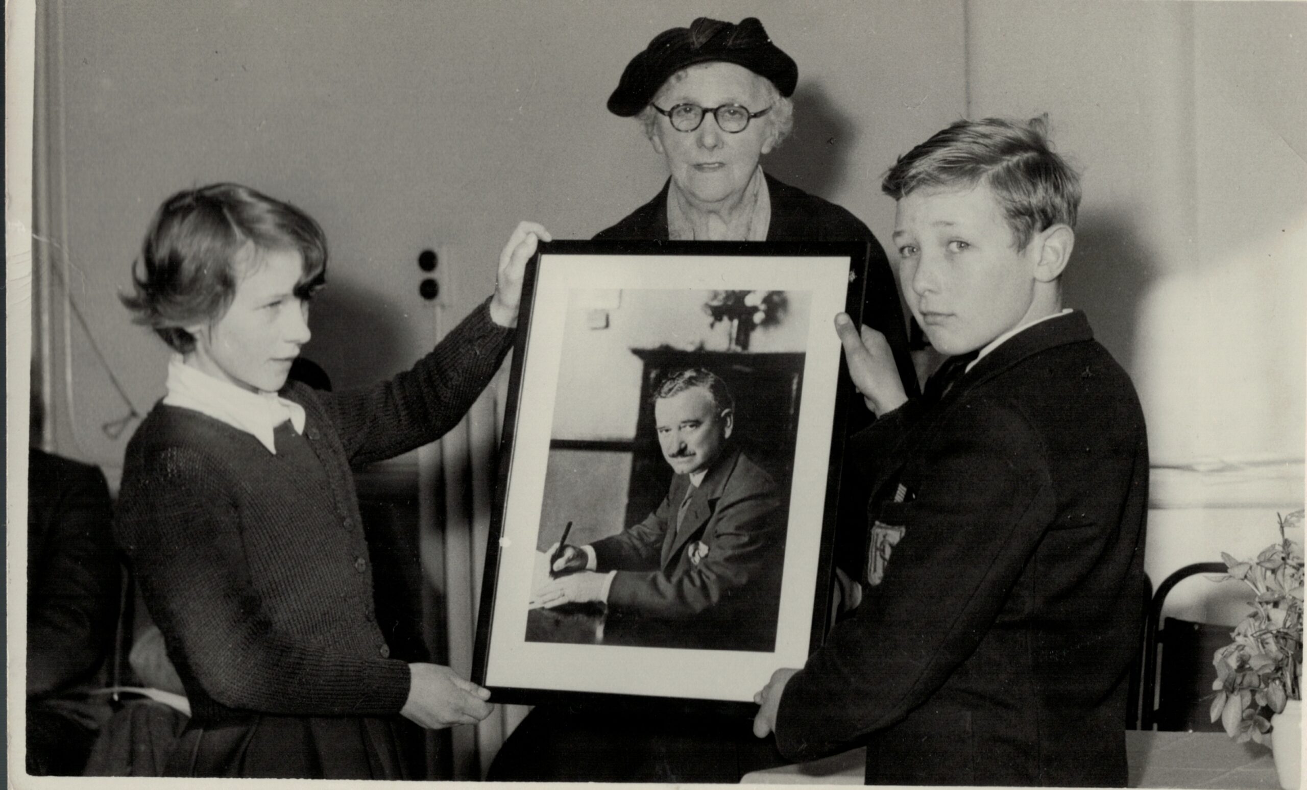 Two school children hold up a large monochrome photo of a man seated at a desk with a pen in his hand. Behind stands a woman in a dark beret and spectacles