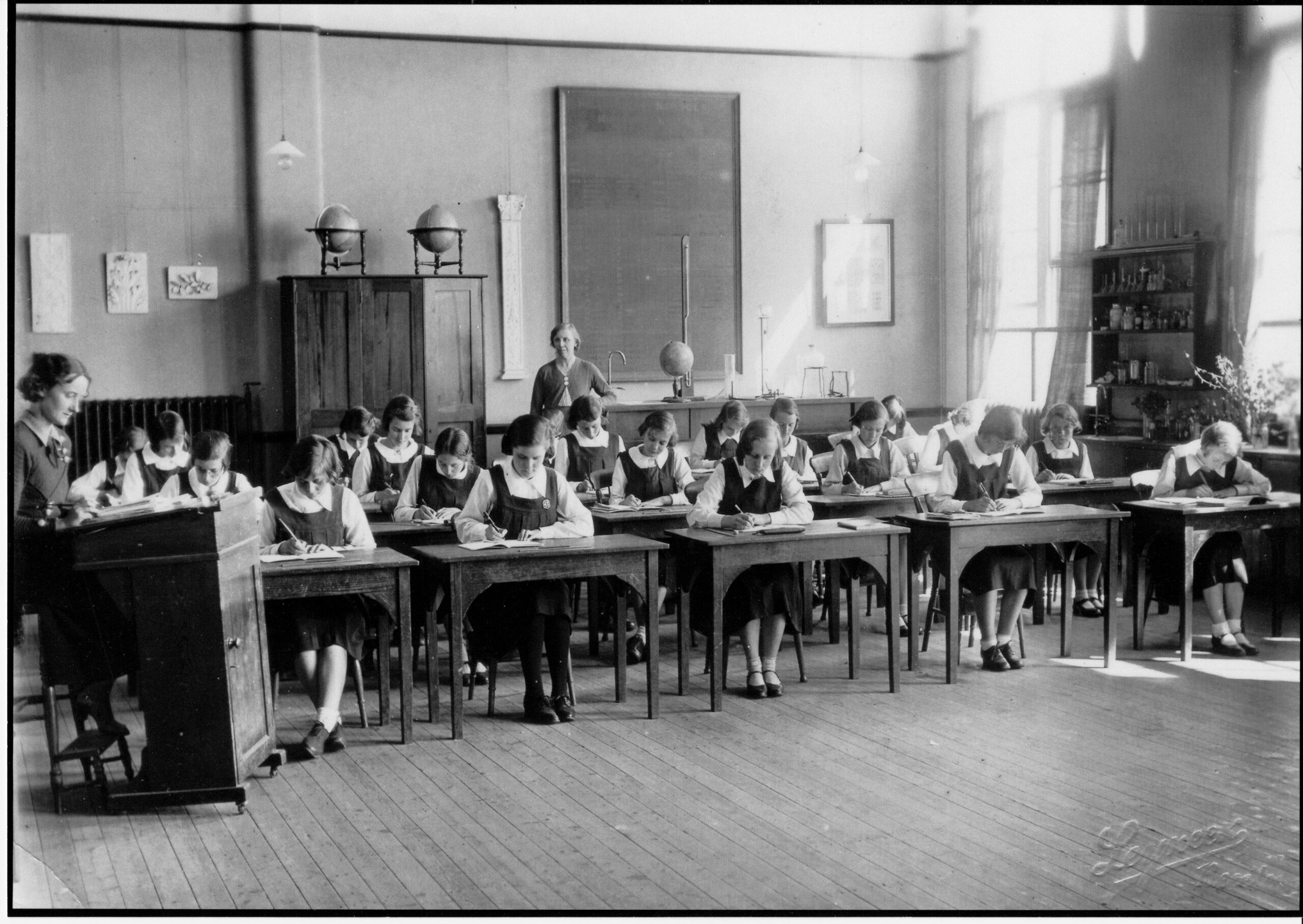 A classroom of girls writing at their desks with members of staff in attendance