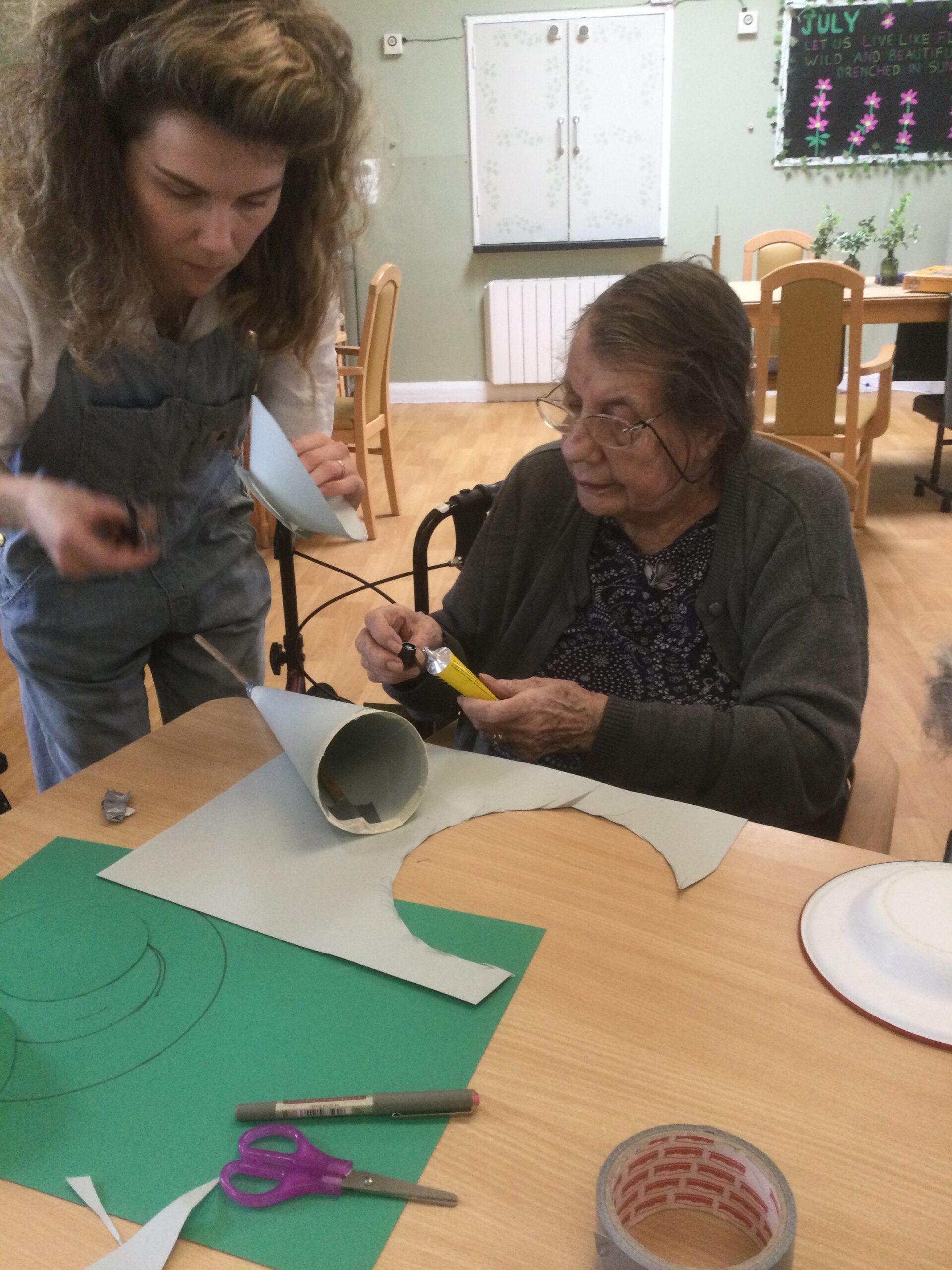 A seated woman replaces the cap on a yellow tube of glue whilst another stands beside her, holding a cardboard dish in her hand