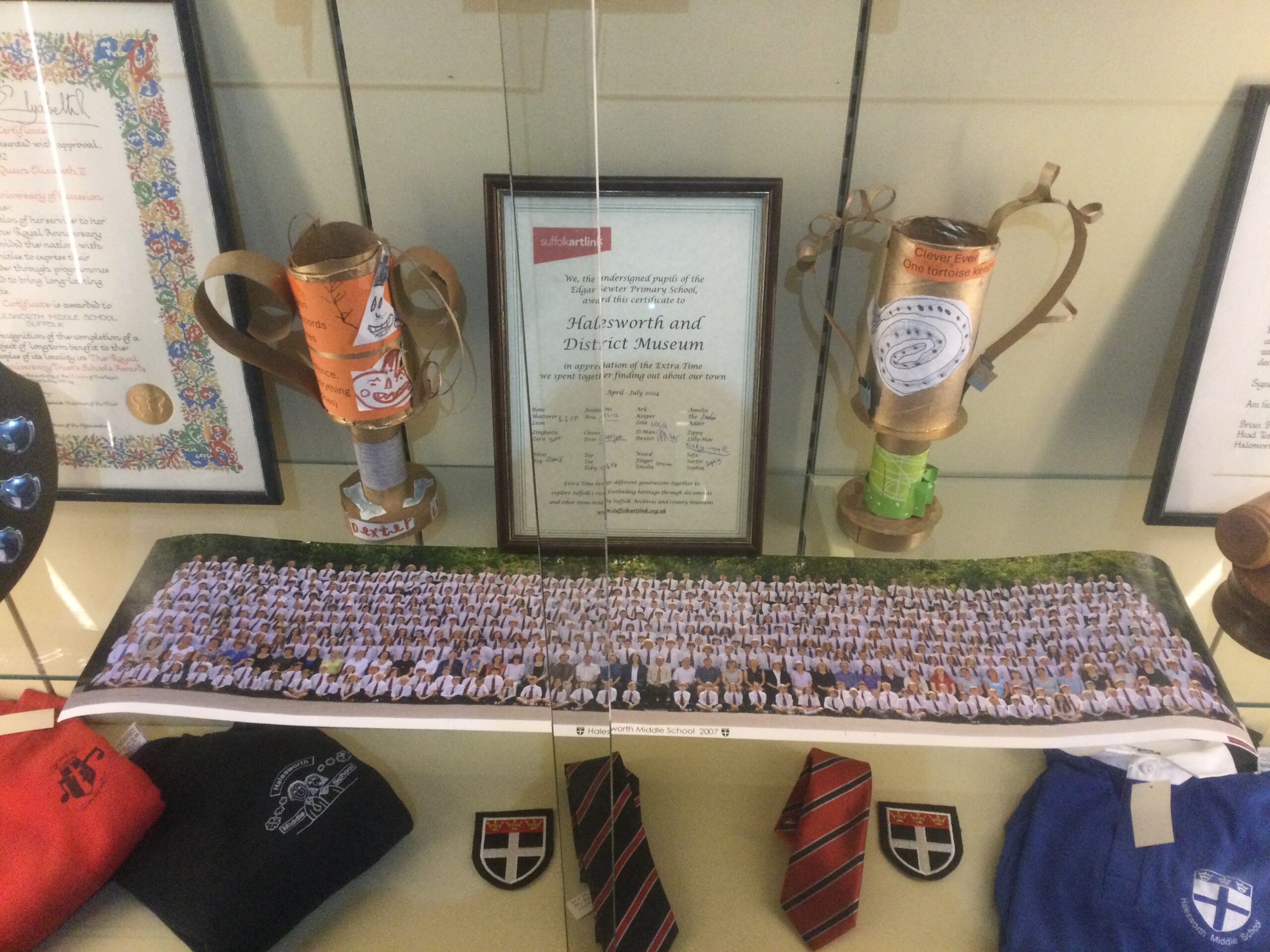 Two hand-crafted trophies stand either side of a framed certificate on a shelf in a display cabinet with a long coloured photograph of all the school children. There are also ties, badges and jumpers in the display