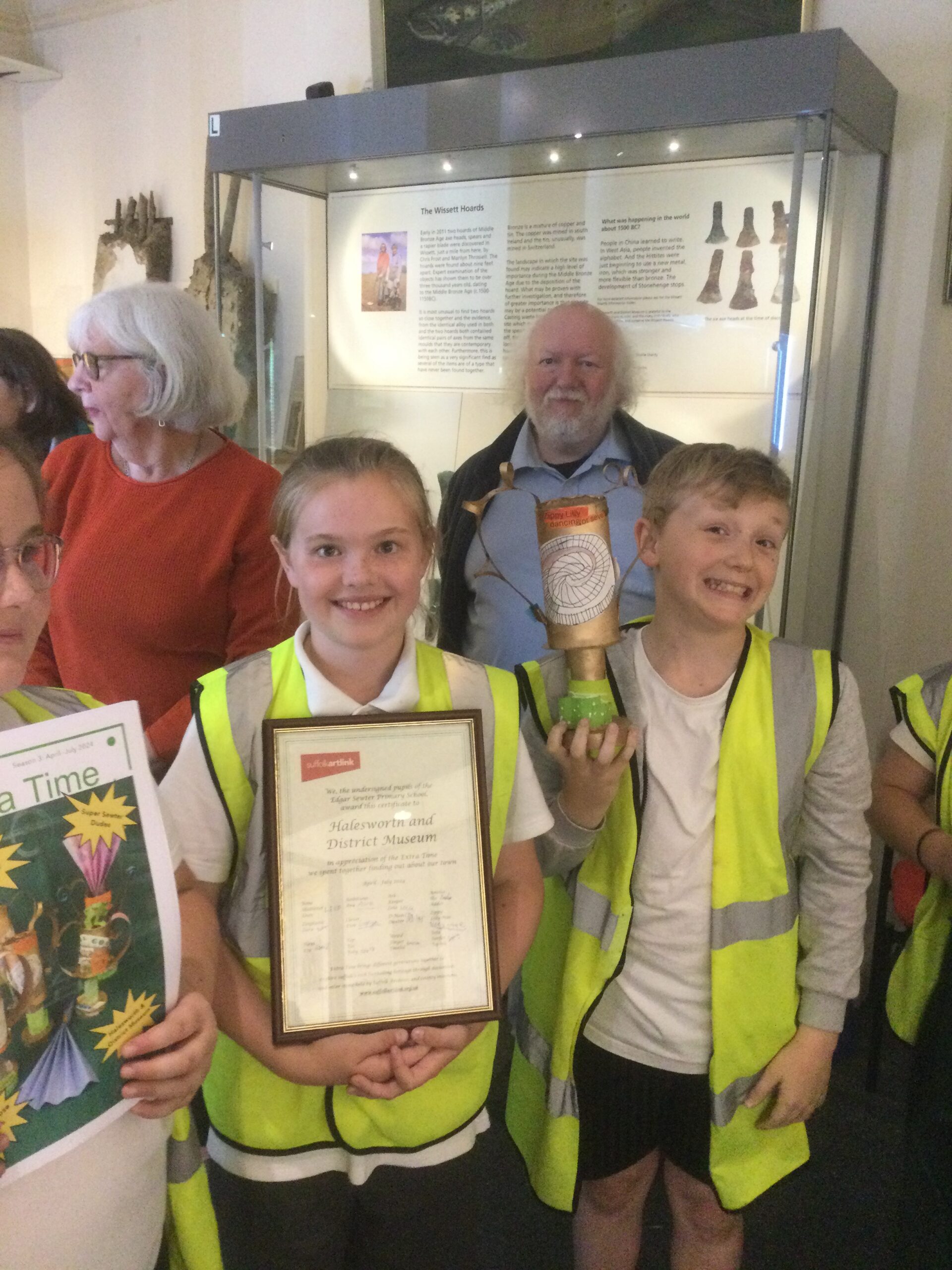 Three children in high vis jackets holding the trophy, certificate and publication. Behind them are two adults - staff at the Museum