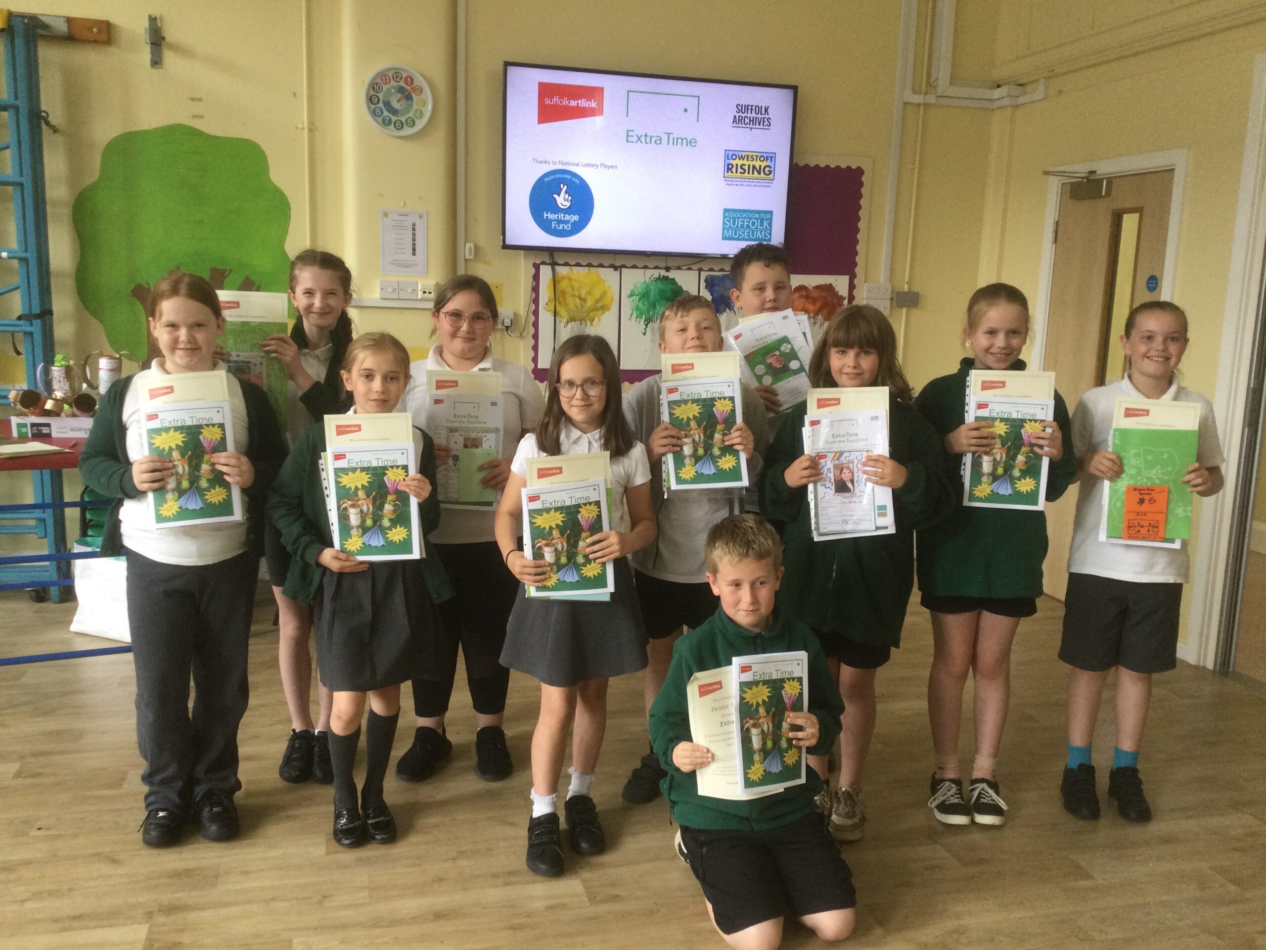 A group of 11 school children stand in the school hall, facing the camera, each holding a certificate and an Extra Time publication. On the wall behind them a large screen carries logos including the National Lottery Heritage Fund logo.