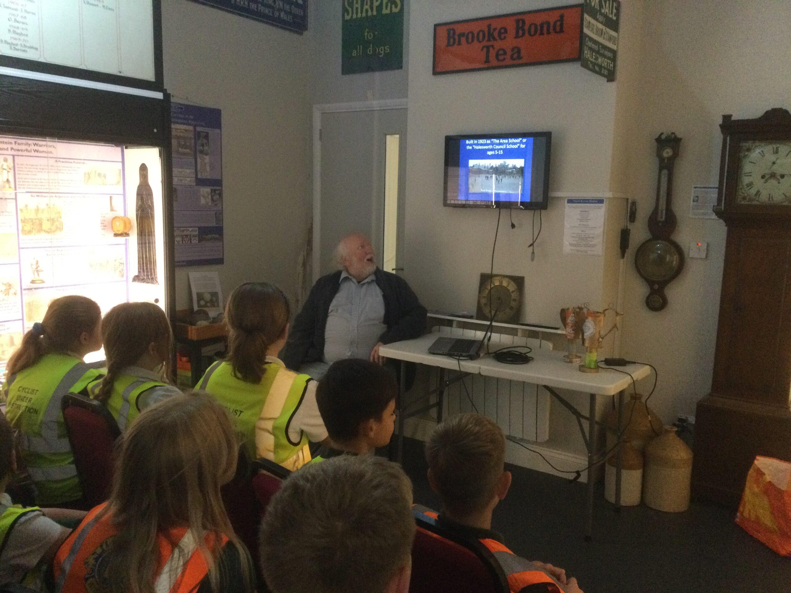 Rows of seated children, all wearing hi-gis jackets, looking at a screen on the wall on which is an old photograph of their school. Other objects in the room include a barometer on the wall, a grandfather clock and an enamel sign Brooke Bond Tea