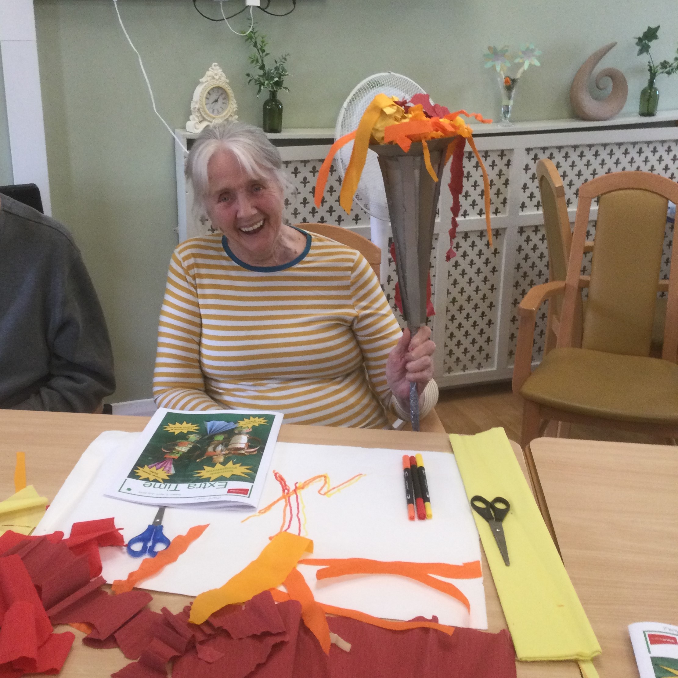 A woman in a yellow and white striped jersey smiles to the camera whilst holding aloft a cardboard Olympic Torch with yellow, orange and red paper 'flames'