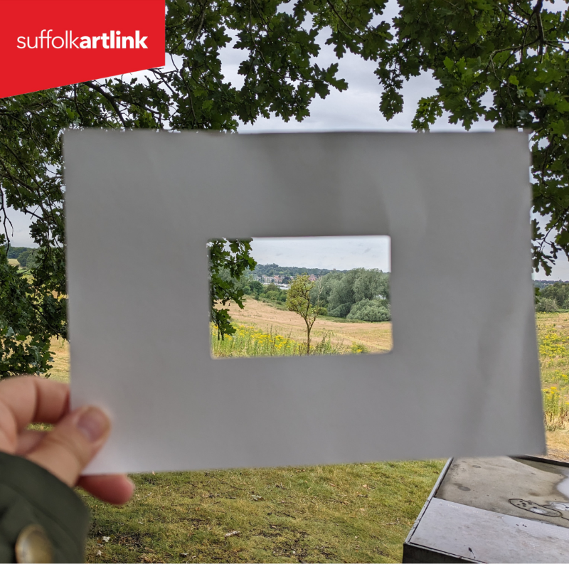 The landscape of Sutton Hoo framed by a handheld rectangular cardboard frame.