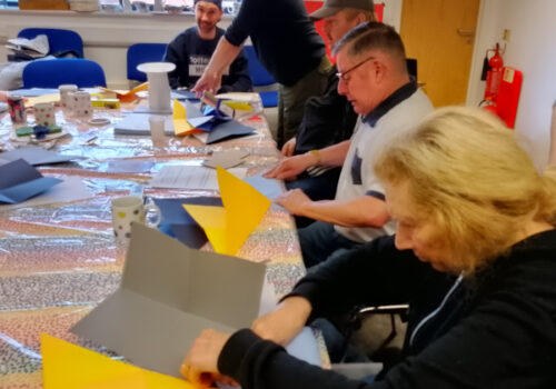 participants of a creative workshop are sitting at a long table. They are intently folding paper to create origami shapes