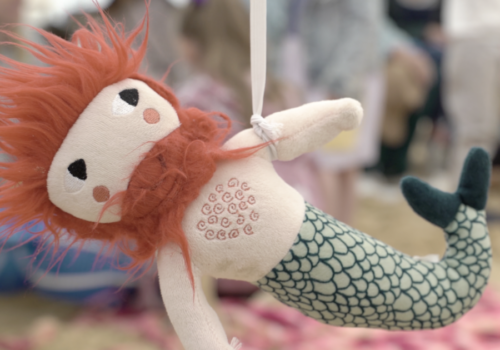 A merman soft toy being dangles by a piece of white ribbon