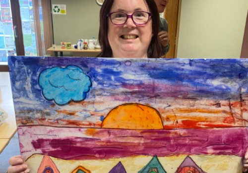 Laura holding her beach painting of meach huts in front of the sea at sunset.