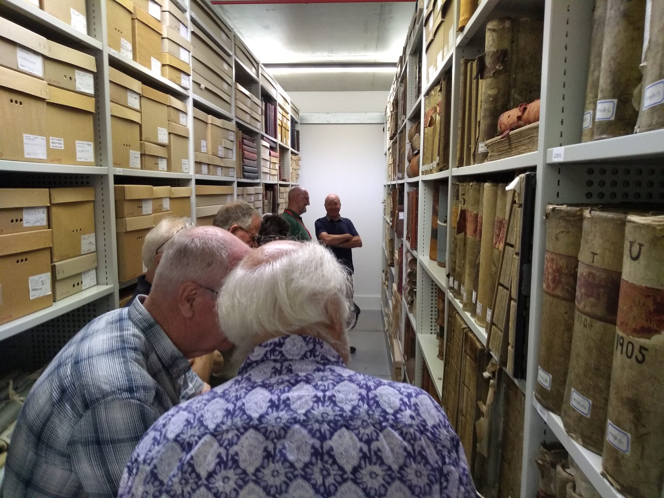 A row of adults in a narrow space between tall sets of metal shelves housing old documents, books and boxes of documents