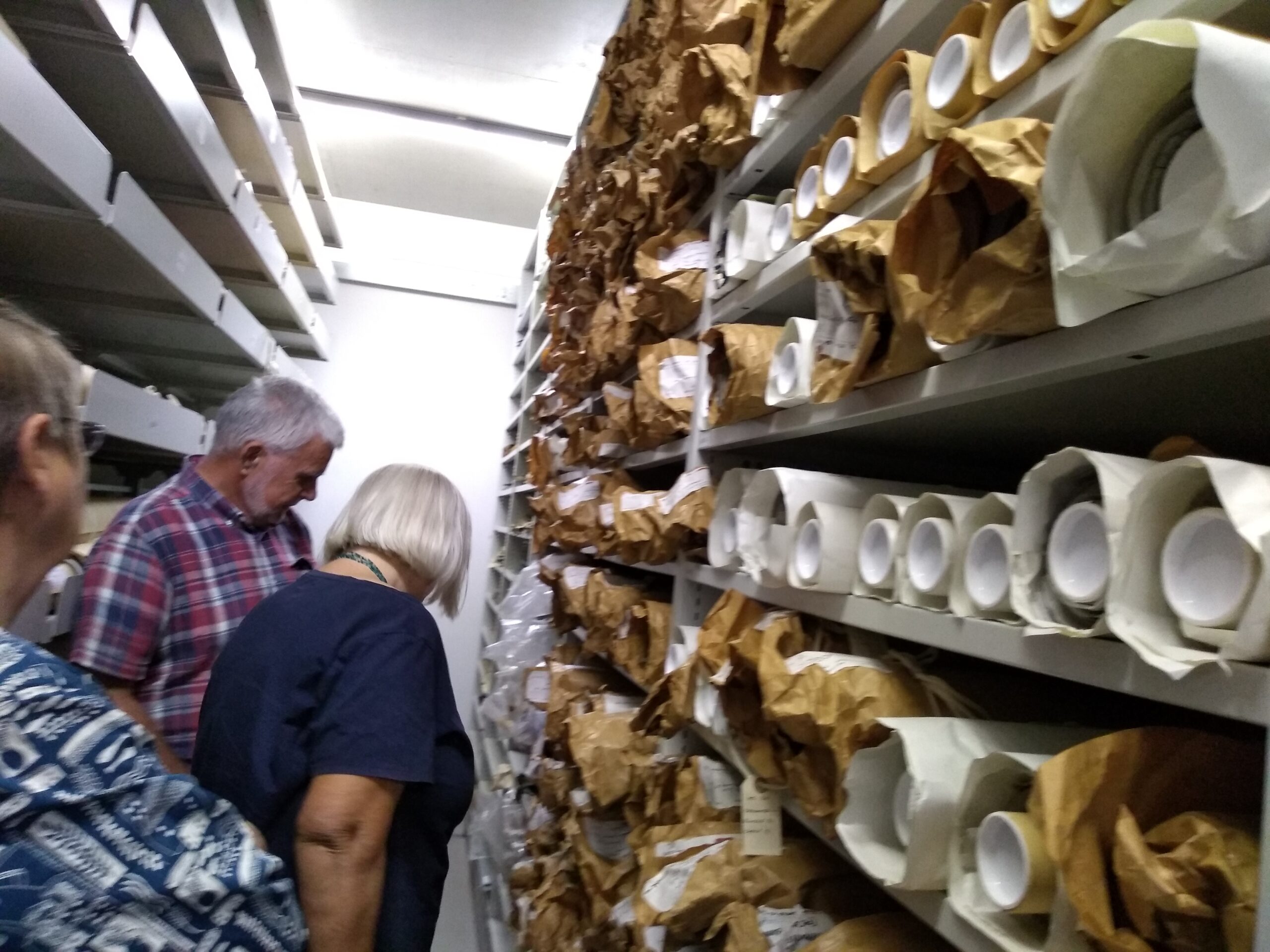 A row of adults in the archive strong room with shelves full of rolled up patterns and drawings, many wrapped in brown paper