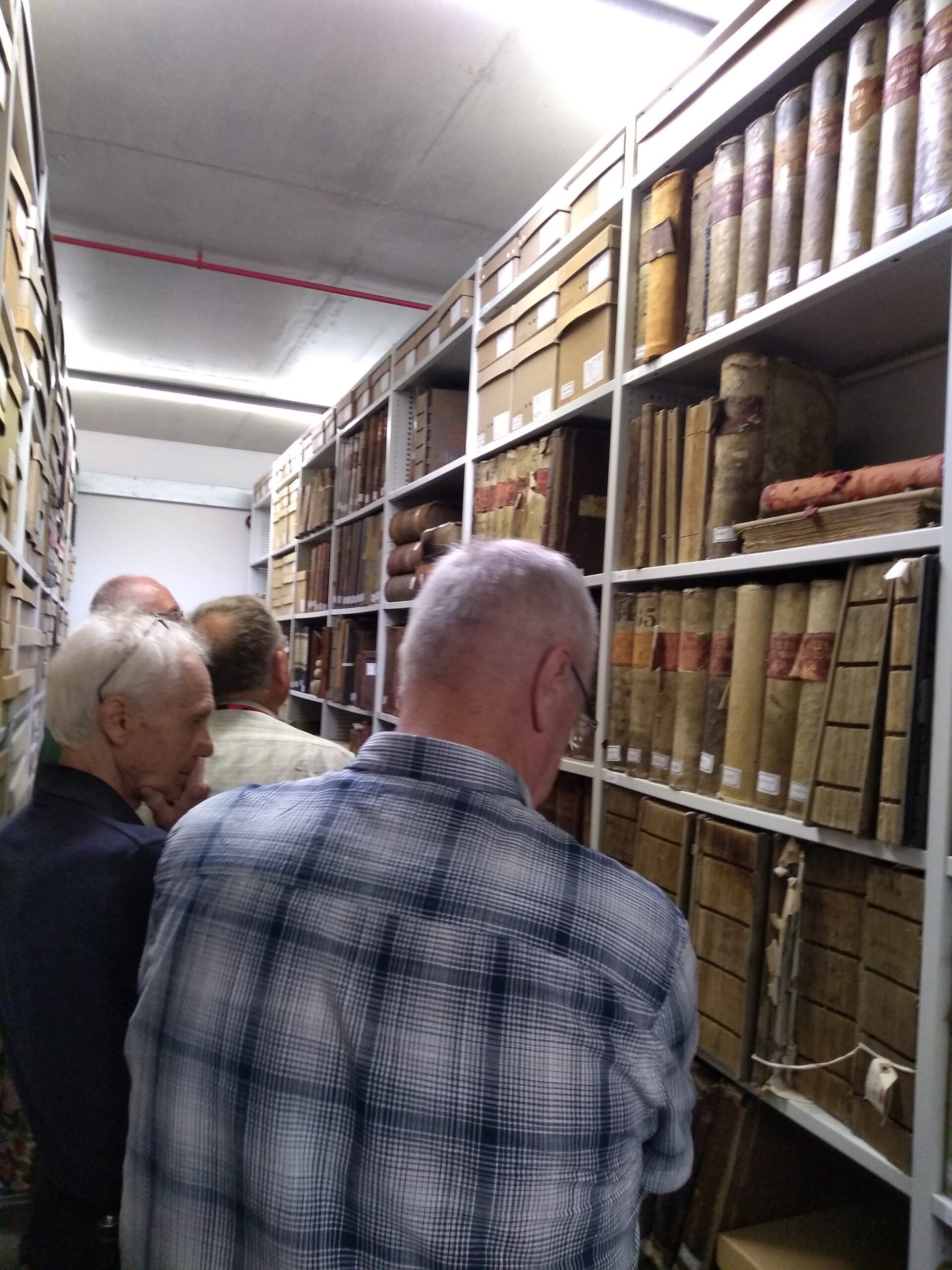 A row of 4 gentlemen in an Archive strongroom with shelves of old books in front and boxes of documents behind