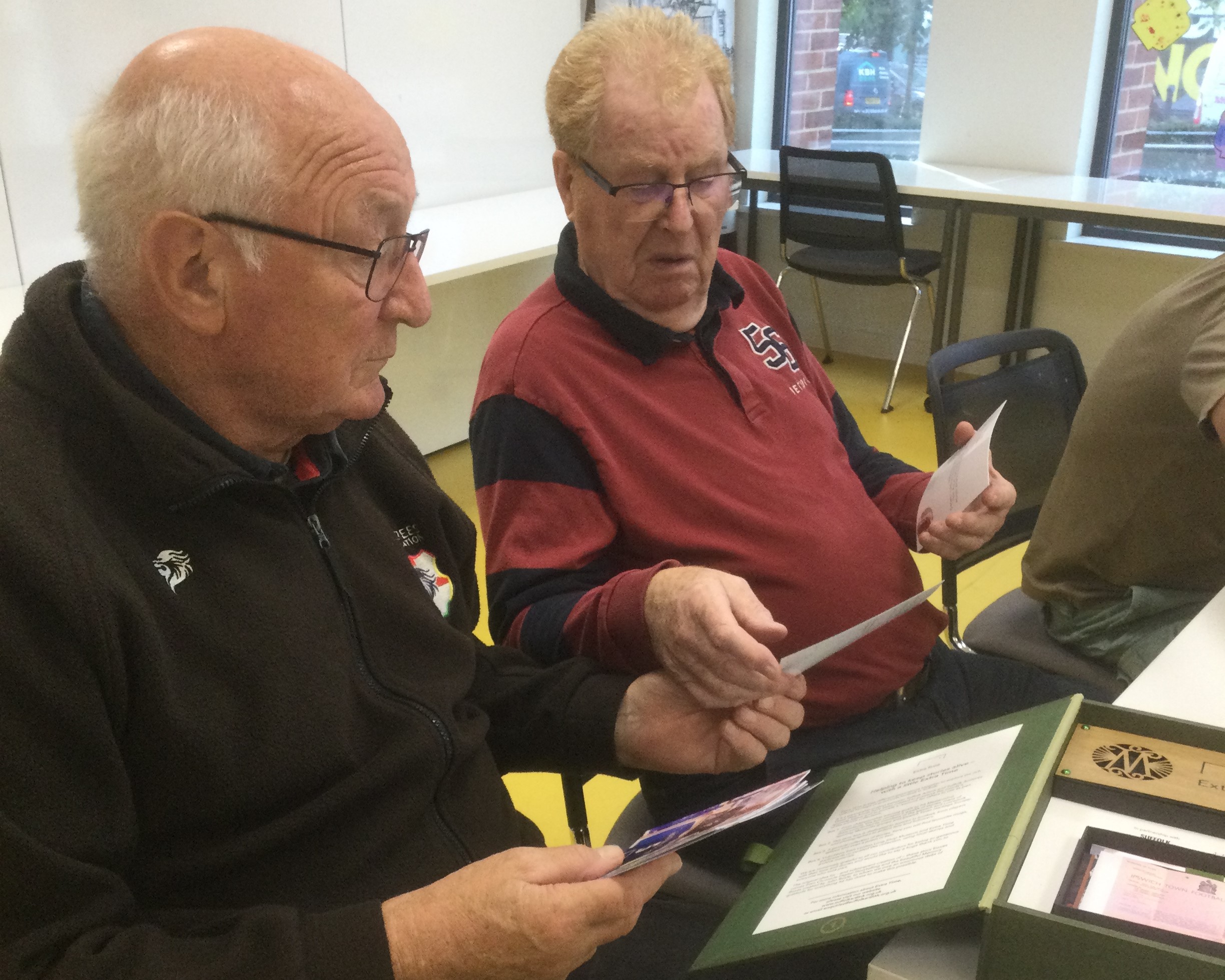 Two gentlemen seated side by side, one holding a pack of photographs and the pair of them discussing them