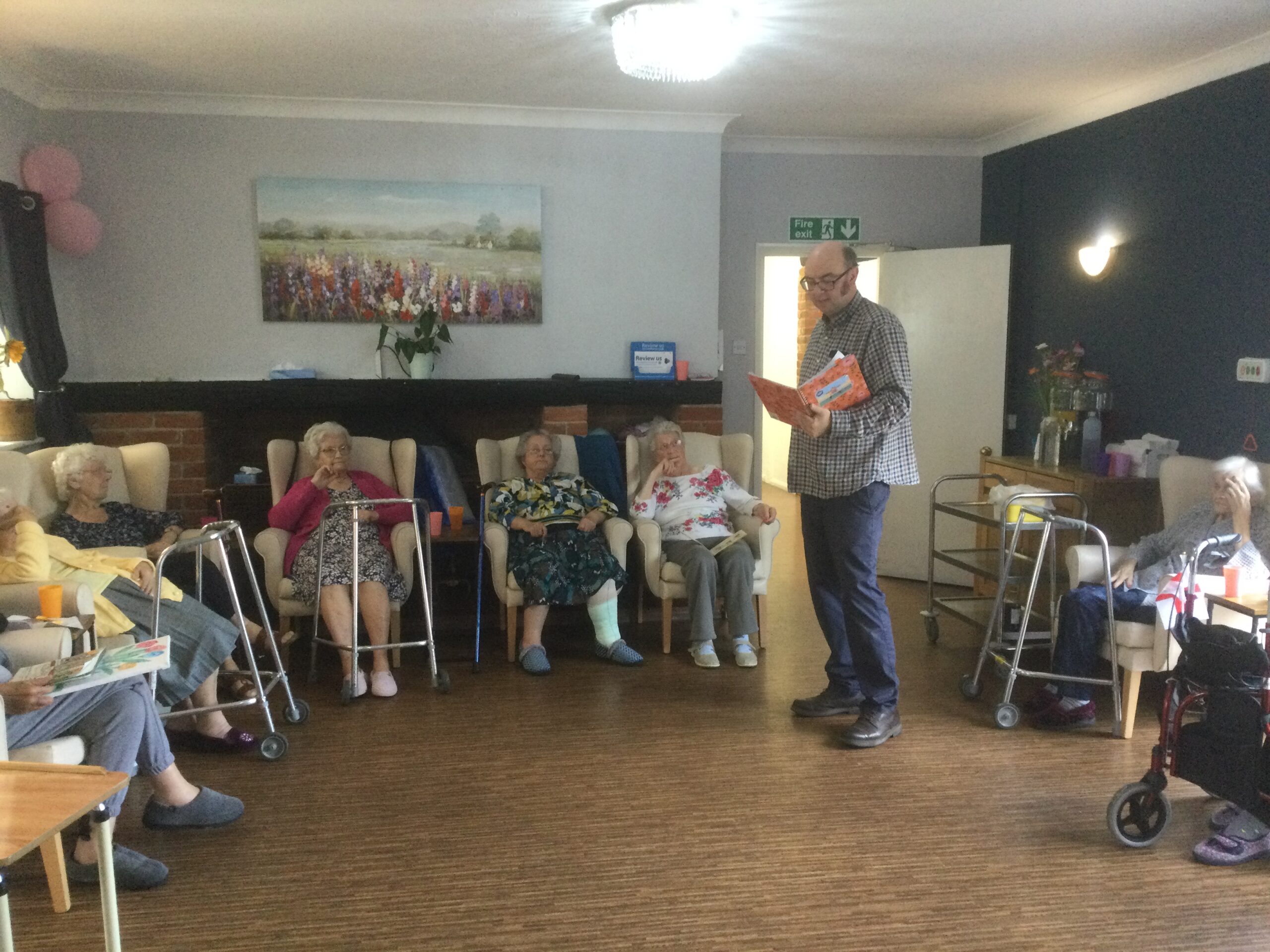 A man stands, reading from a large book whilst around the room are seated several ladies, some of them with Zimmer frames in front of them. There's a large picture on the back wall and two pink balloons in the corner on the wall, left