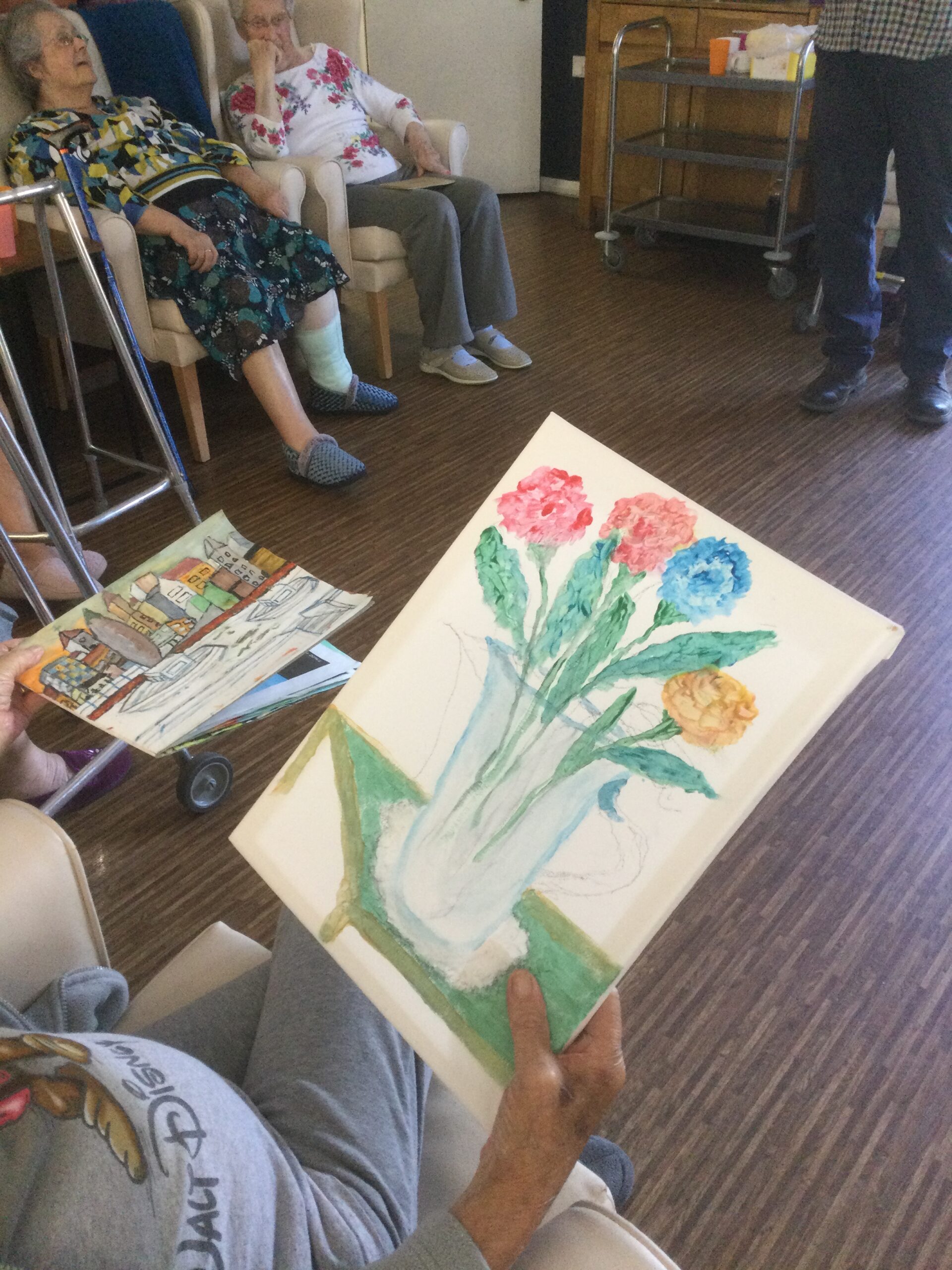 A view over a person's shoulder, showing him holding a white canvas with a painting of four flowers in a vase.