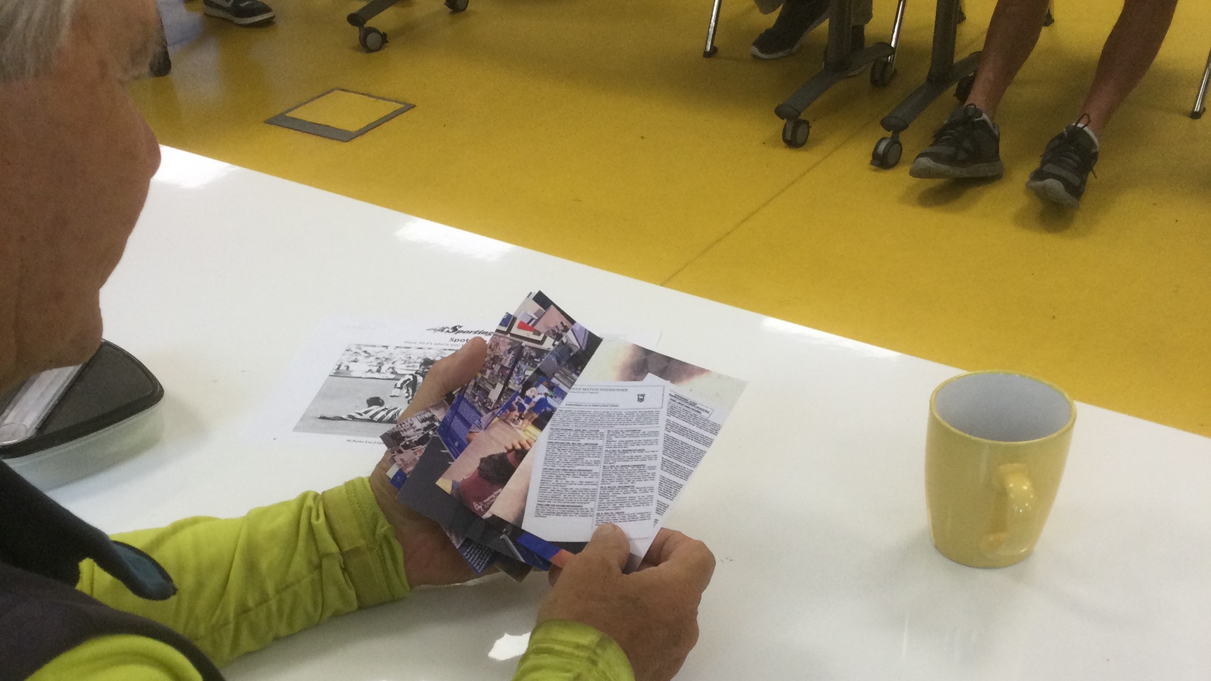 A person seated at a white table, a yellow coffee mug to one side. In his hand is a pack of photographs.