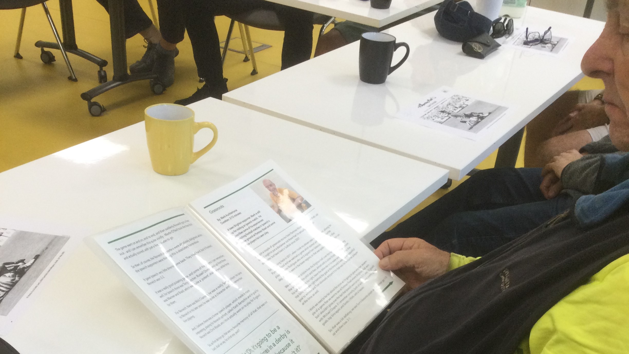 A person seated at a table, with a booklet in his hands. On the table are coffee mugs and pieces of paper.