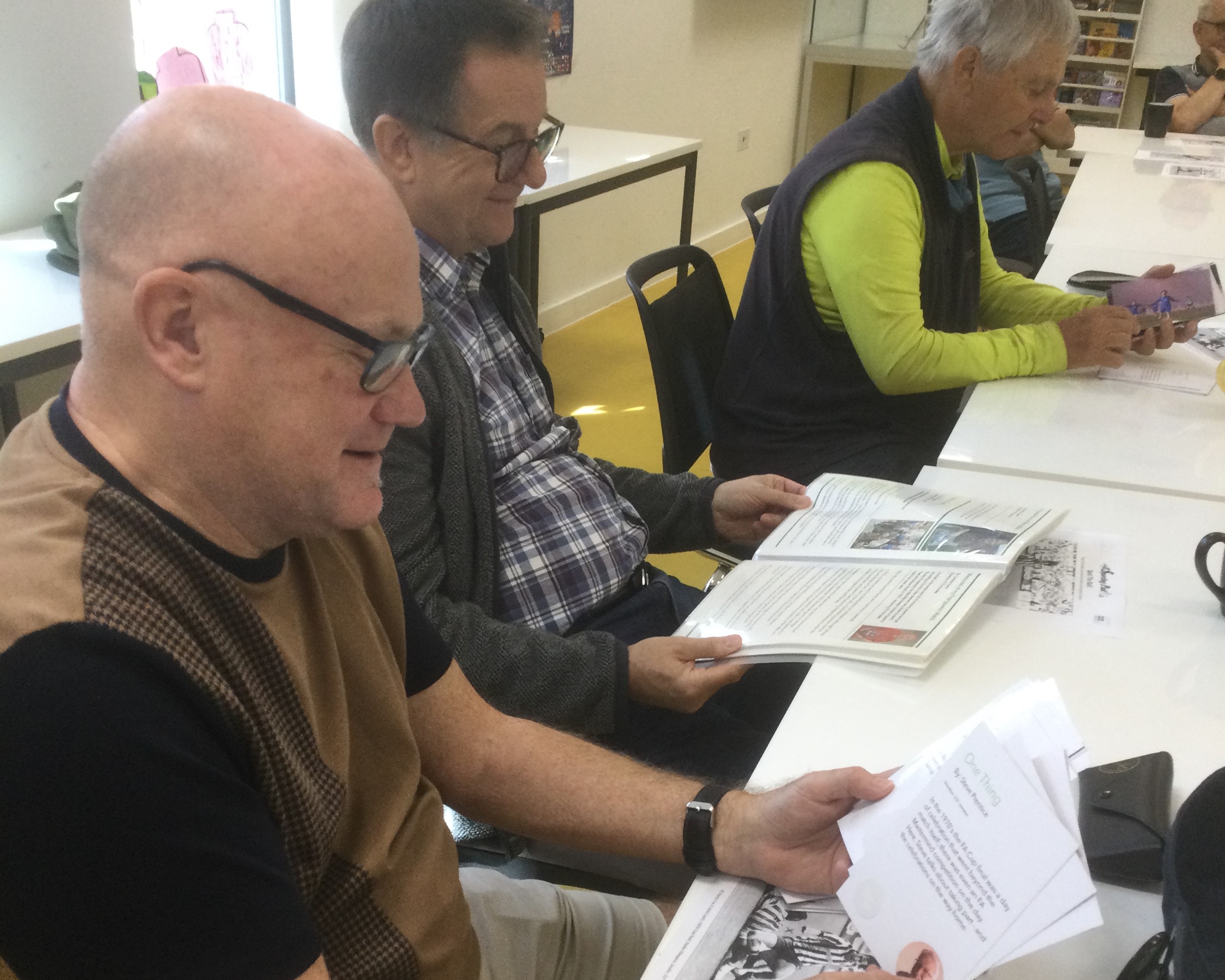 Three men seated at white tables, each looking at photographs and stories about footballing and all smiling.