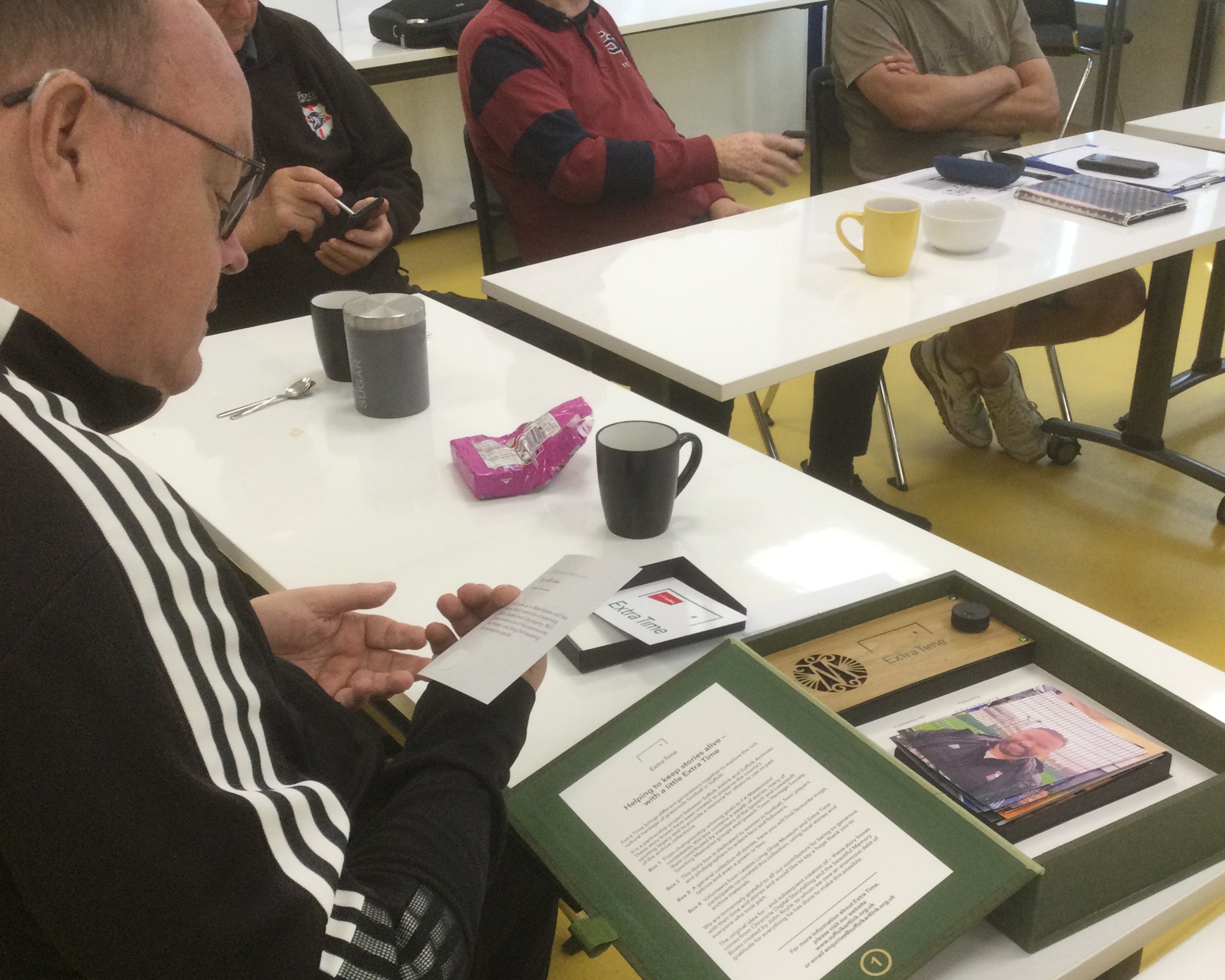 A man reads the back of a card. In front is an open green box with more photographs inside. Other people are seated at tables along from him, with coffee cups and a packet of biscuits.