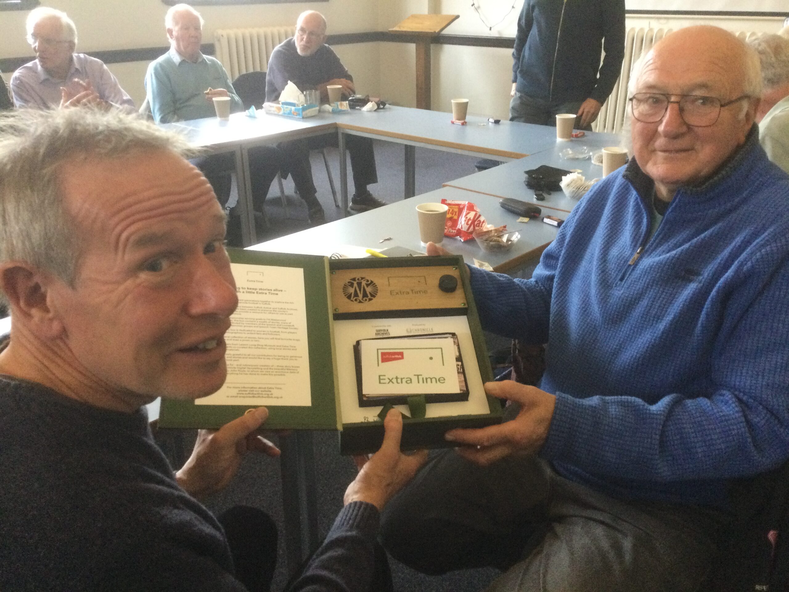 Two gentlemen in the foreground seated at a blue table and holding a green box file that has a small speaker panel and photographs. In the background are three gentlemen chatting amongst themselves