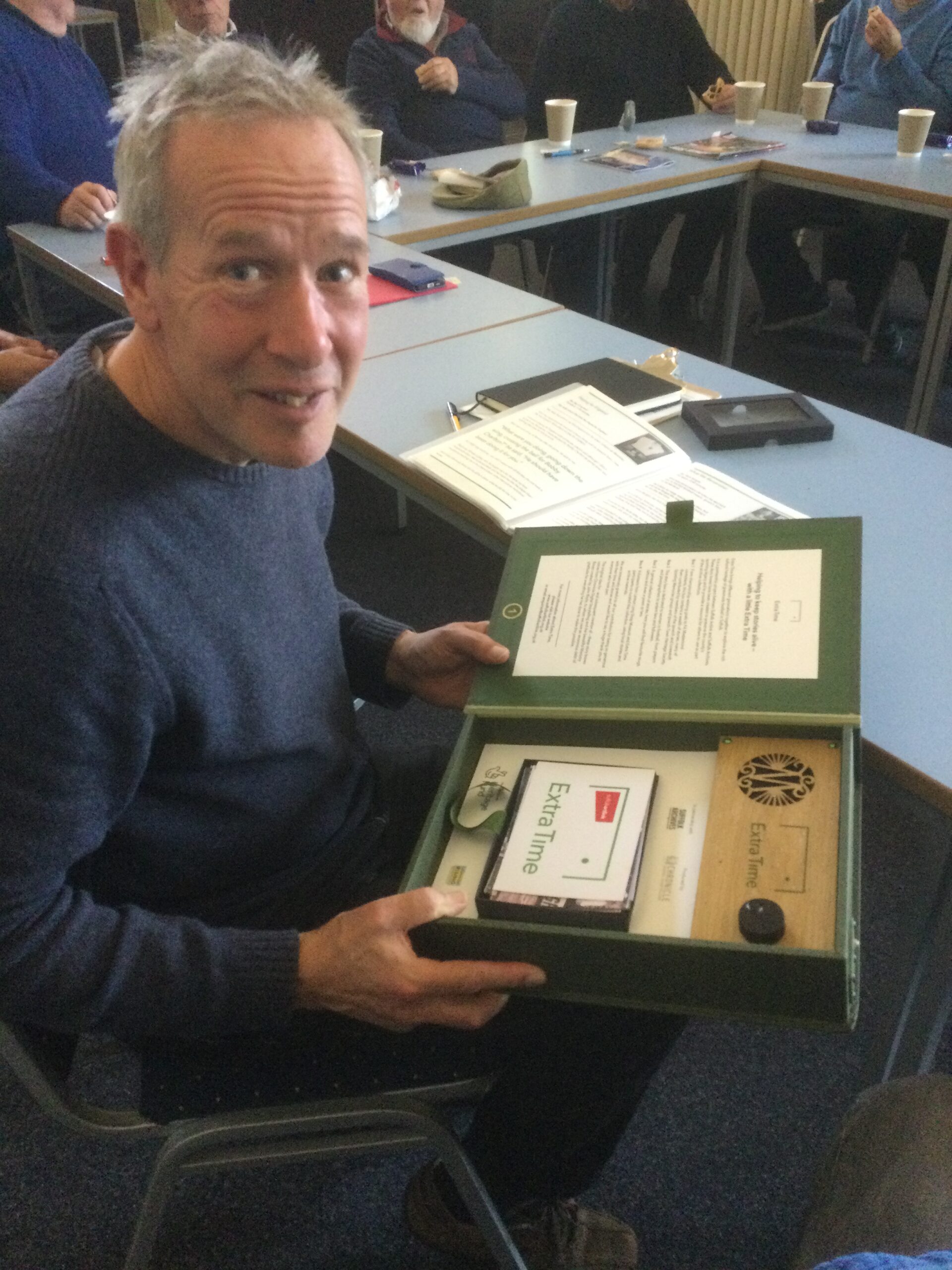 A gentleman in a blue jersey seated at a row of blue tables, holding a green box file