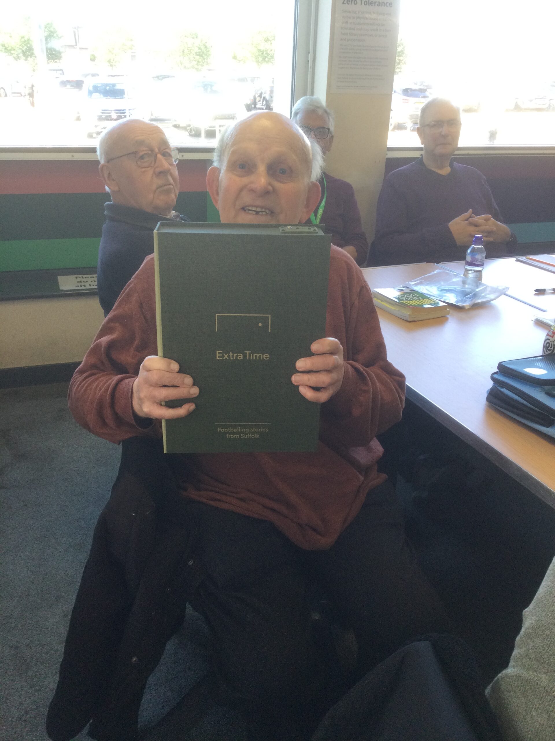 A gentleman holds the green box file to his chest, facing camera whilst other men are seated in the background, looking on