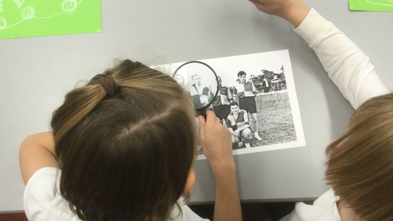 Overhead view of two children, one with a magnifying glass in hand, studying a black and white photograph of a football team