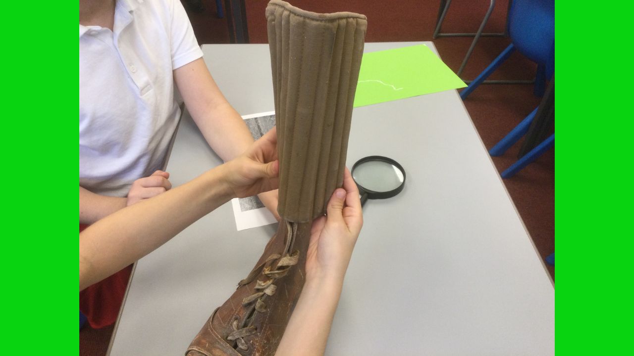 A pair of hands outstretched, holding an old shin pad on top of an old leather football boot whilst a second pair of hands steadies the boot