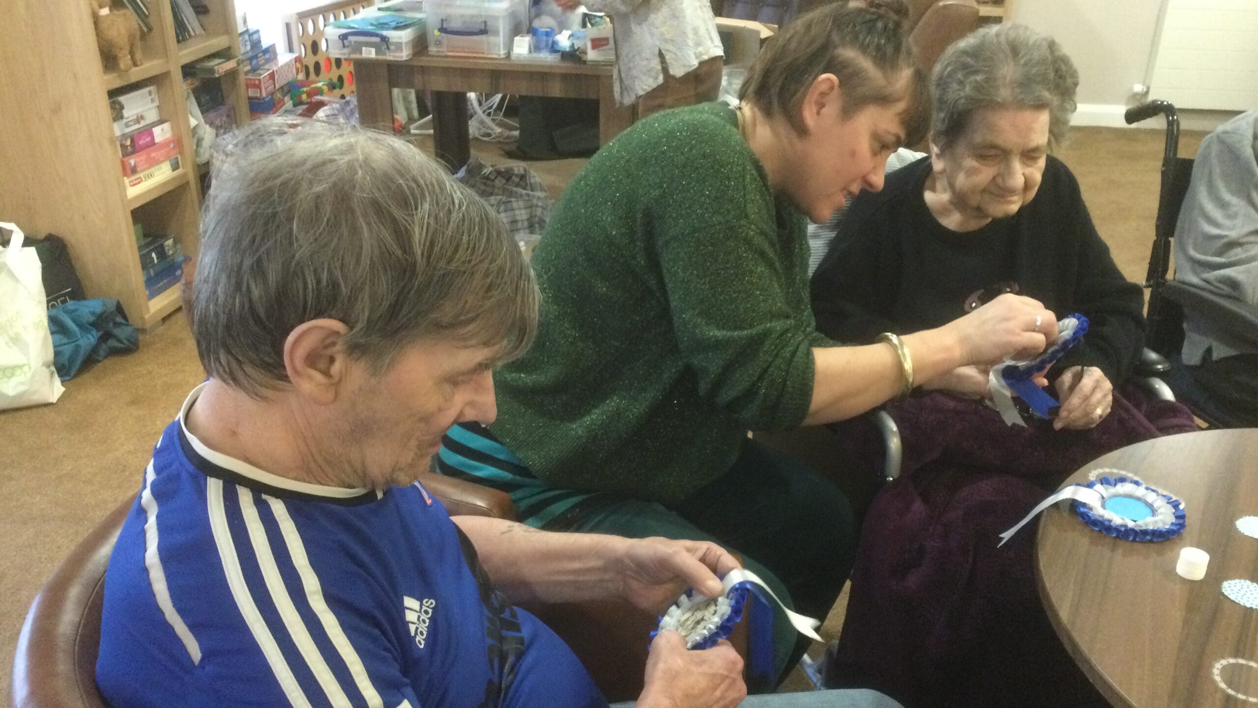 A person in a green jumper leans towards an elderly lady, wearing black, and together they are looking at something held by the woman. In the foreground, a man wearing a blue and white football top holds a rosette in his hands